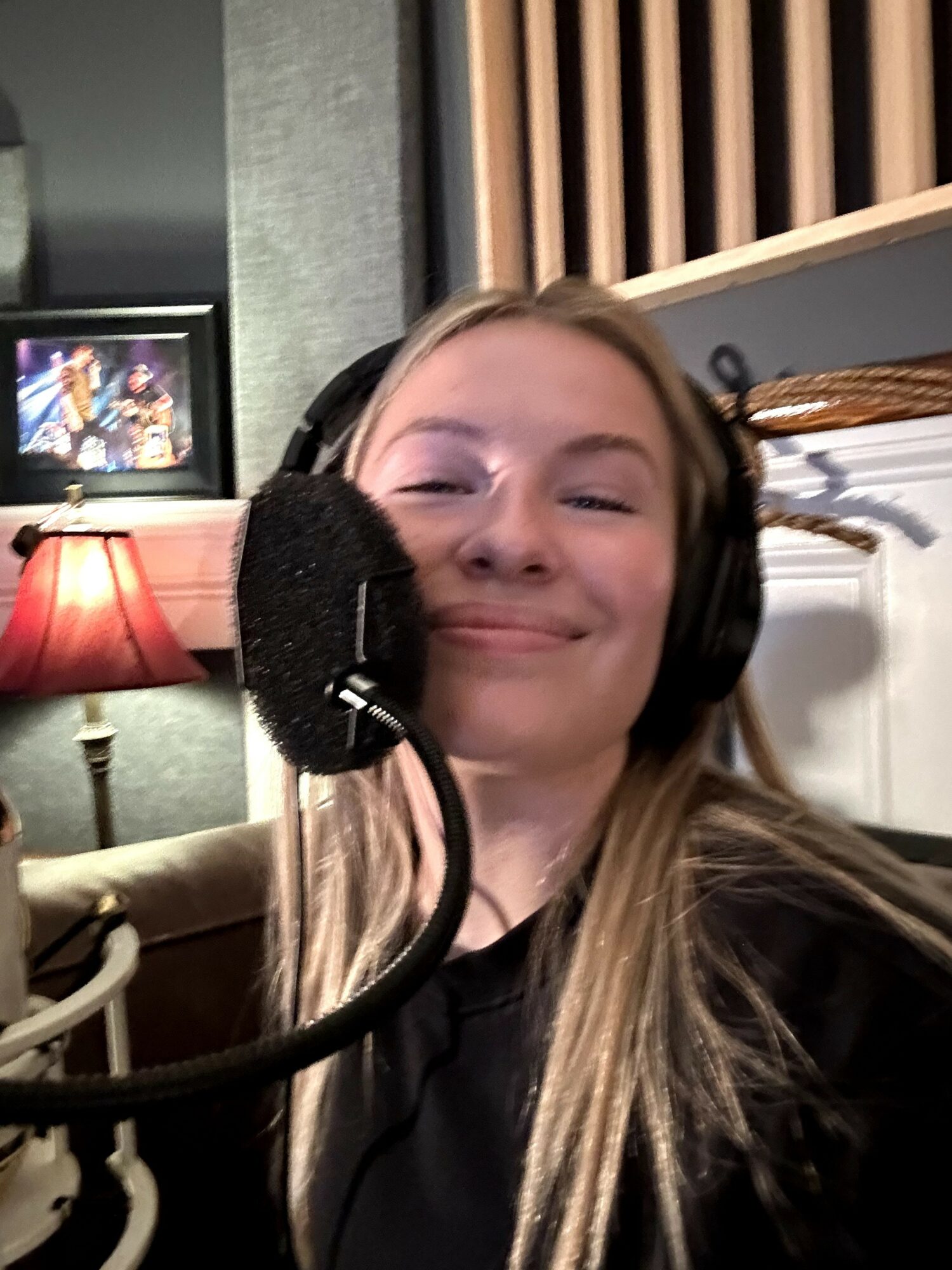 Young woman with long hair wearing headphones, smiling, in a recording studio with a microphone in front.