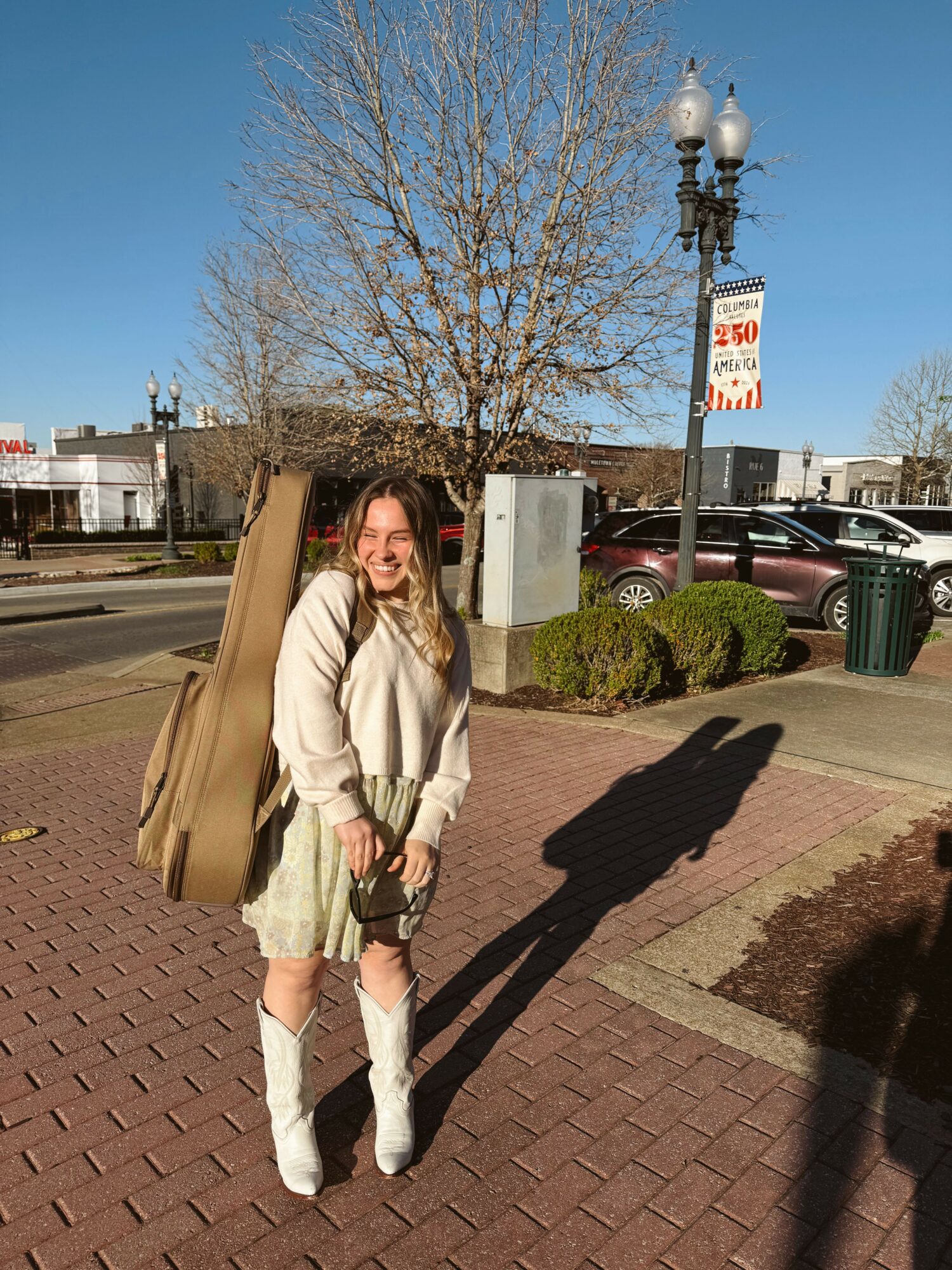 Woman standing on brick sidewalk with shadow, trees, and street scene in background.
