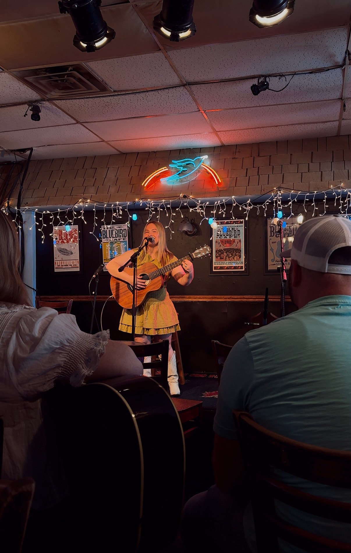 Person playing guitar on stage with audience watching, decorated with string lights and posters, neon sign above.