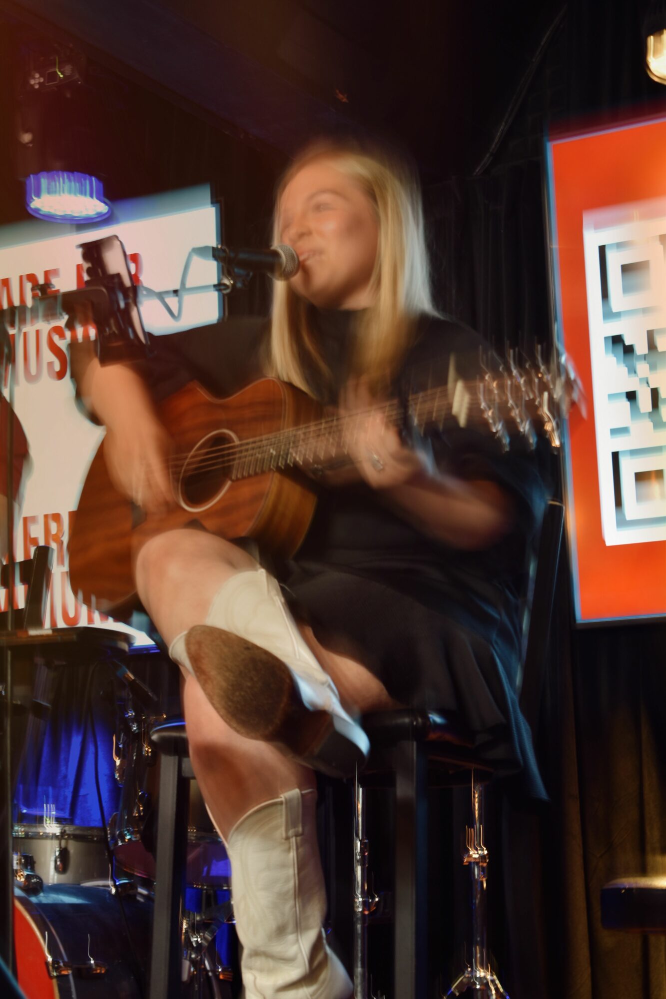 Person playing guitar on stage with musical equipment and a screen in the background.