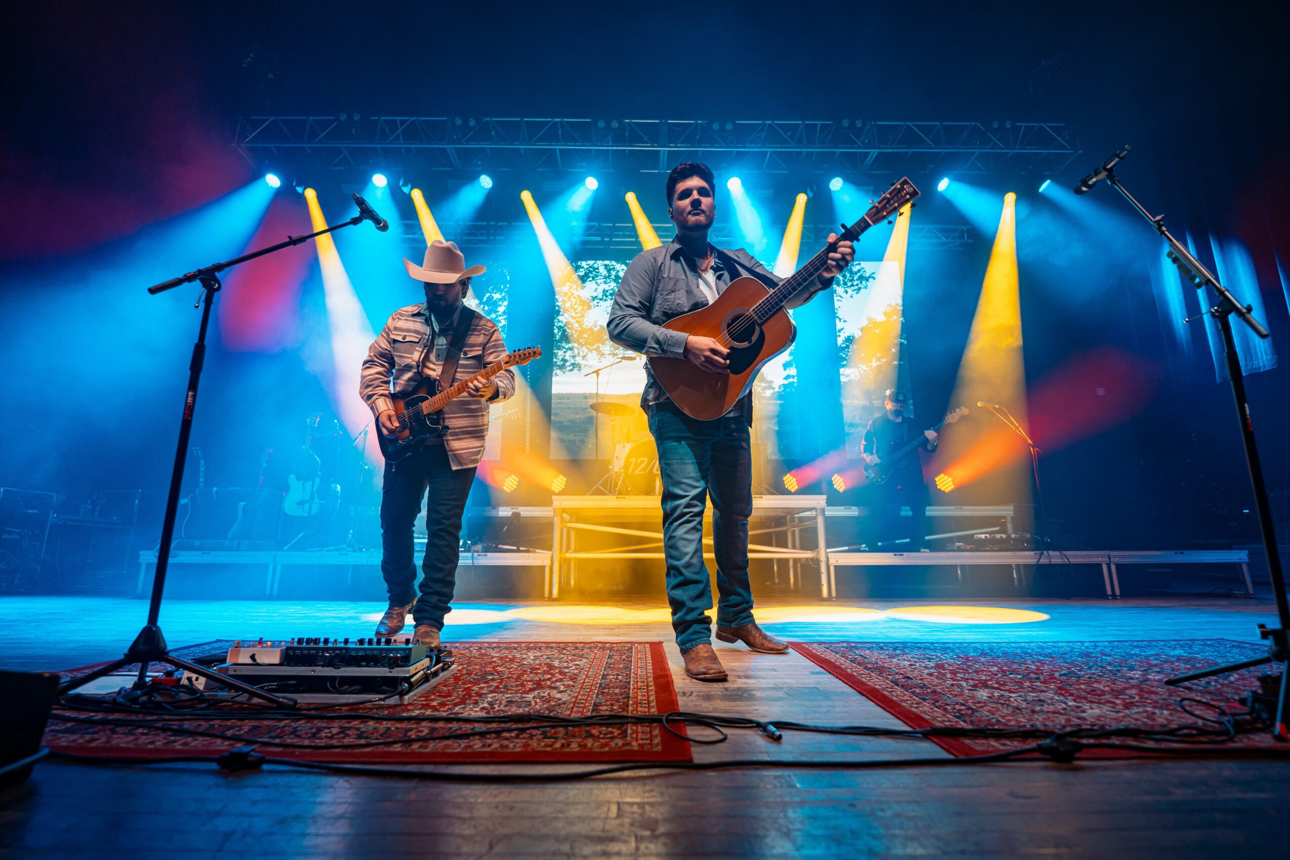 Two musicians perform on stage with colorful lighting and a backdrop, one playing guitar and the other wearing a hat and playing an instrument.