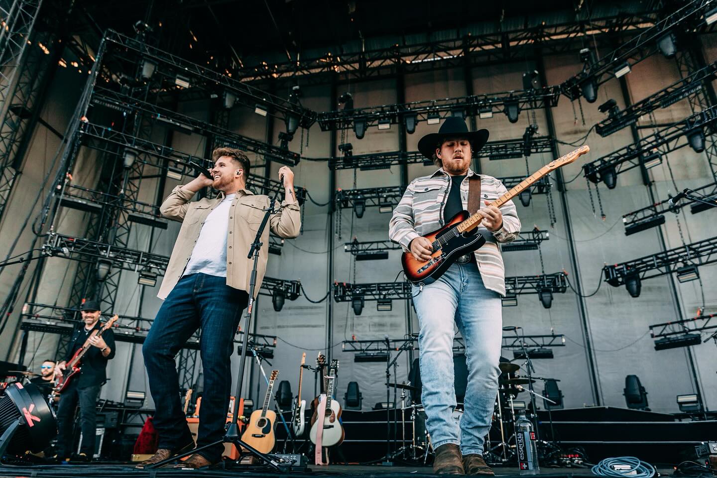 Two musicians perform on stage with guitars, one singing into a microphone, stage equipment and additional guitars visible behind them.