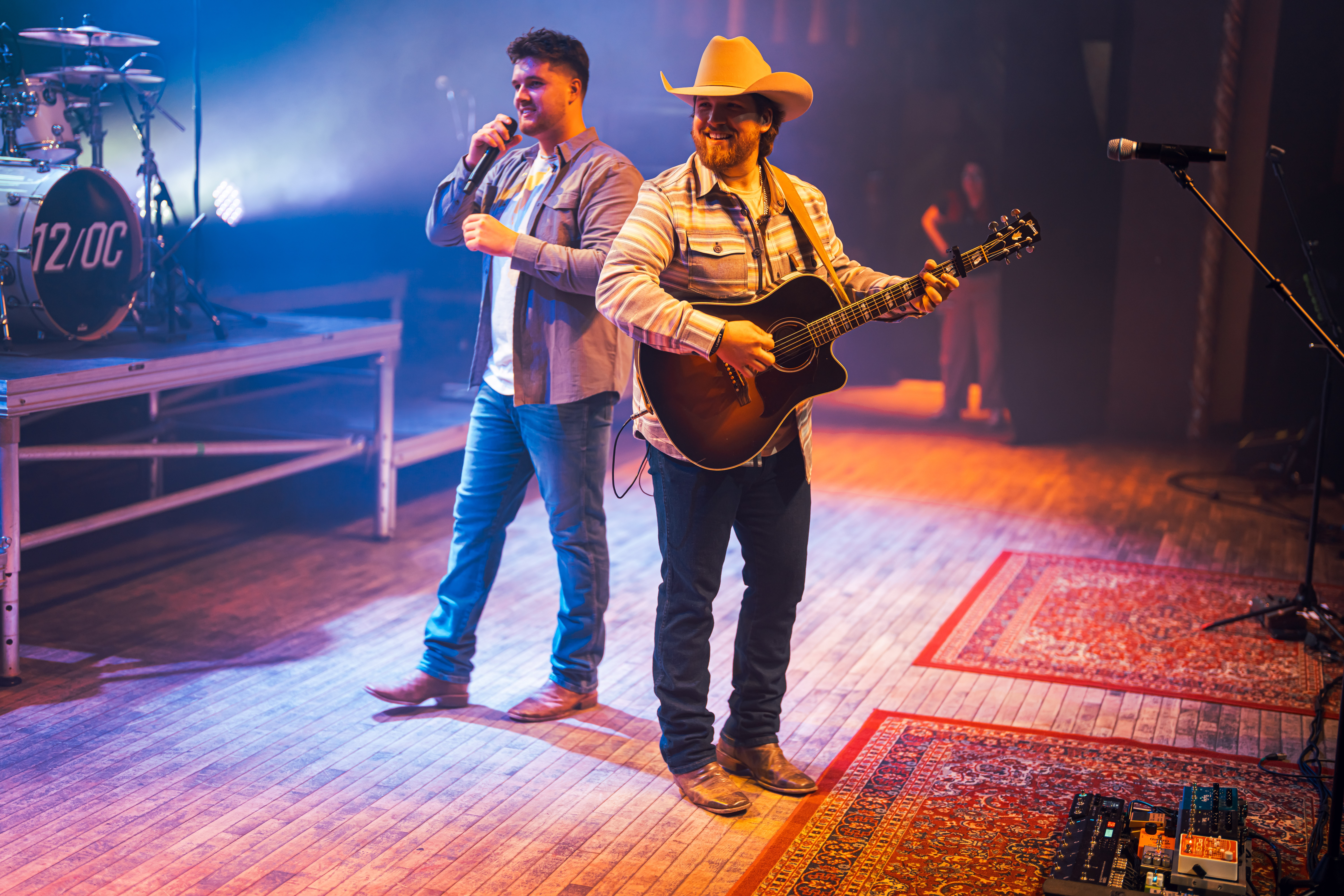 Two musicians perform on stage, one singing into a microphone and the other playing guitar, with stage lighting and equipment visible.