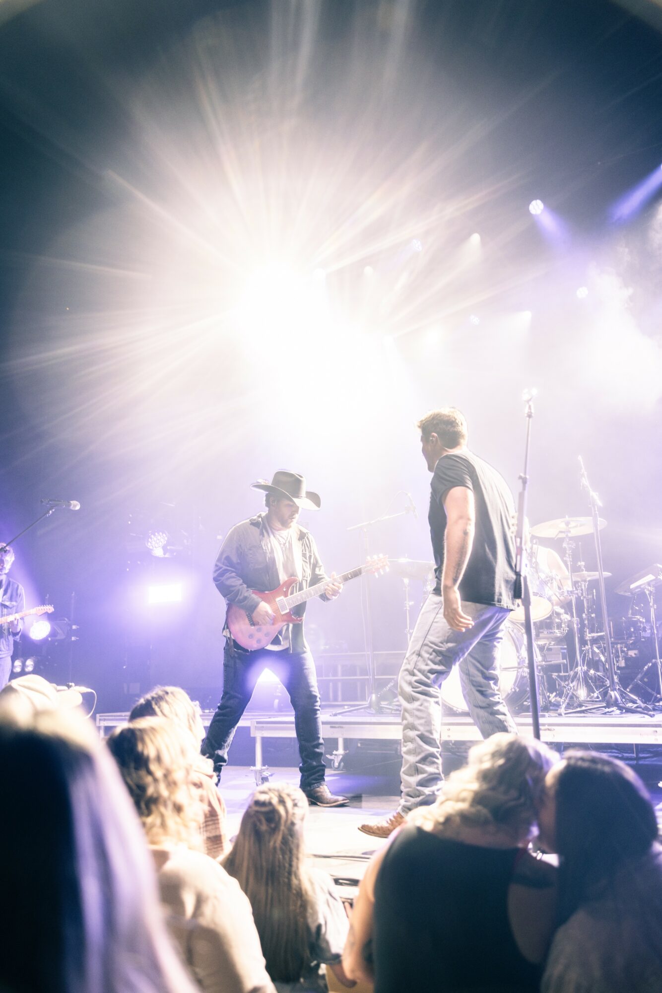 Two musicians perform on stage with bright lights and fog, audience visible in foreground.