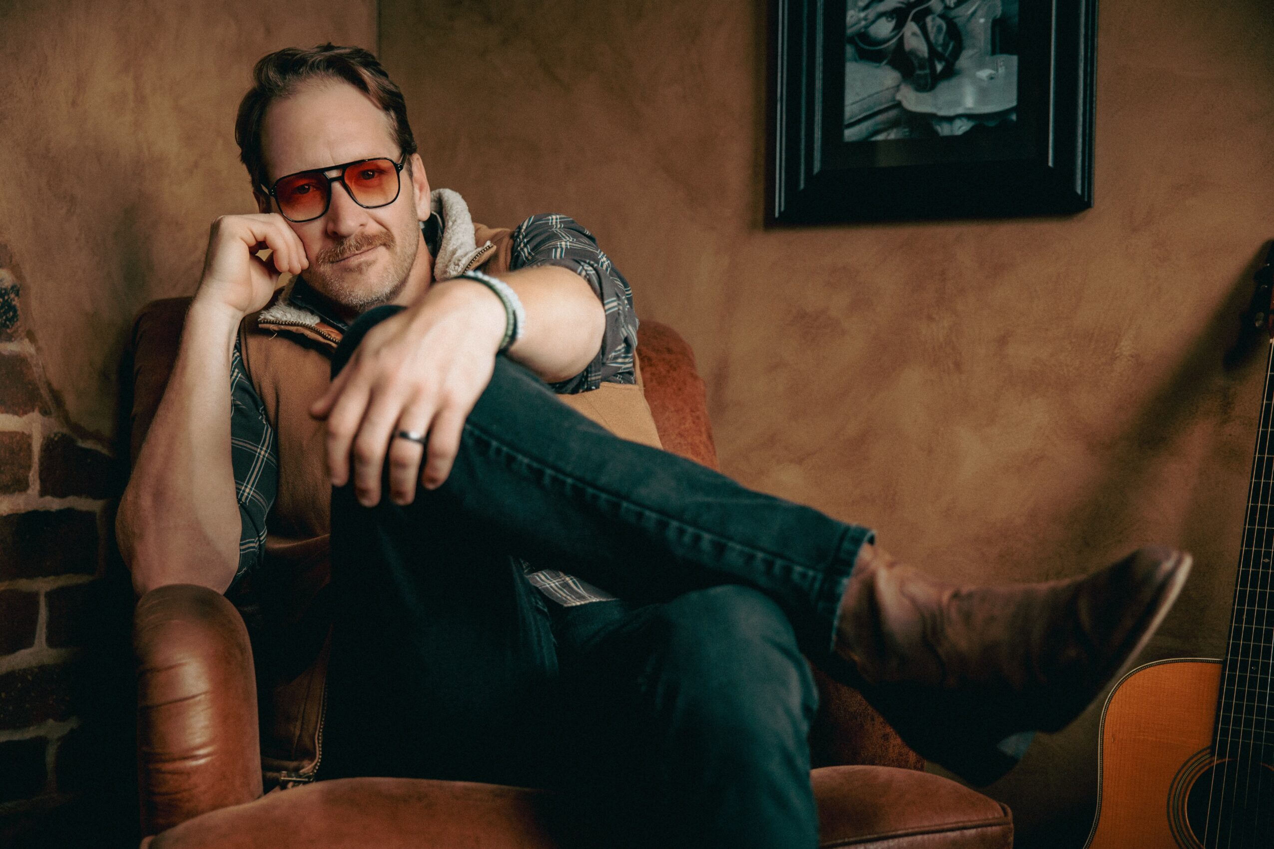 Man with glasses and headphones sitting on a leather chair, smiling, with a guitar nearby, in a cozy room.
