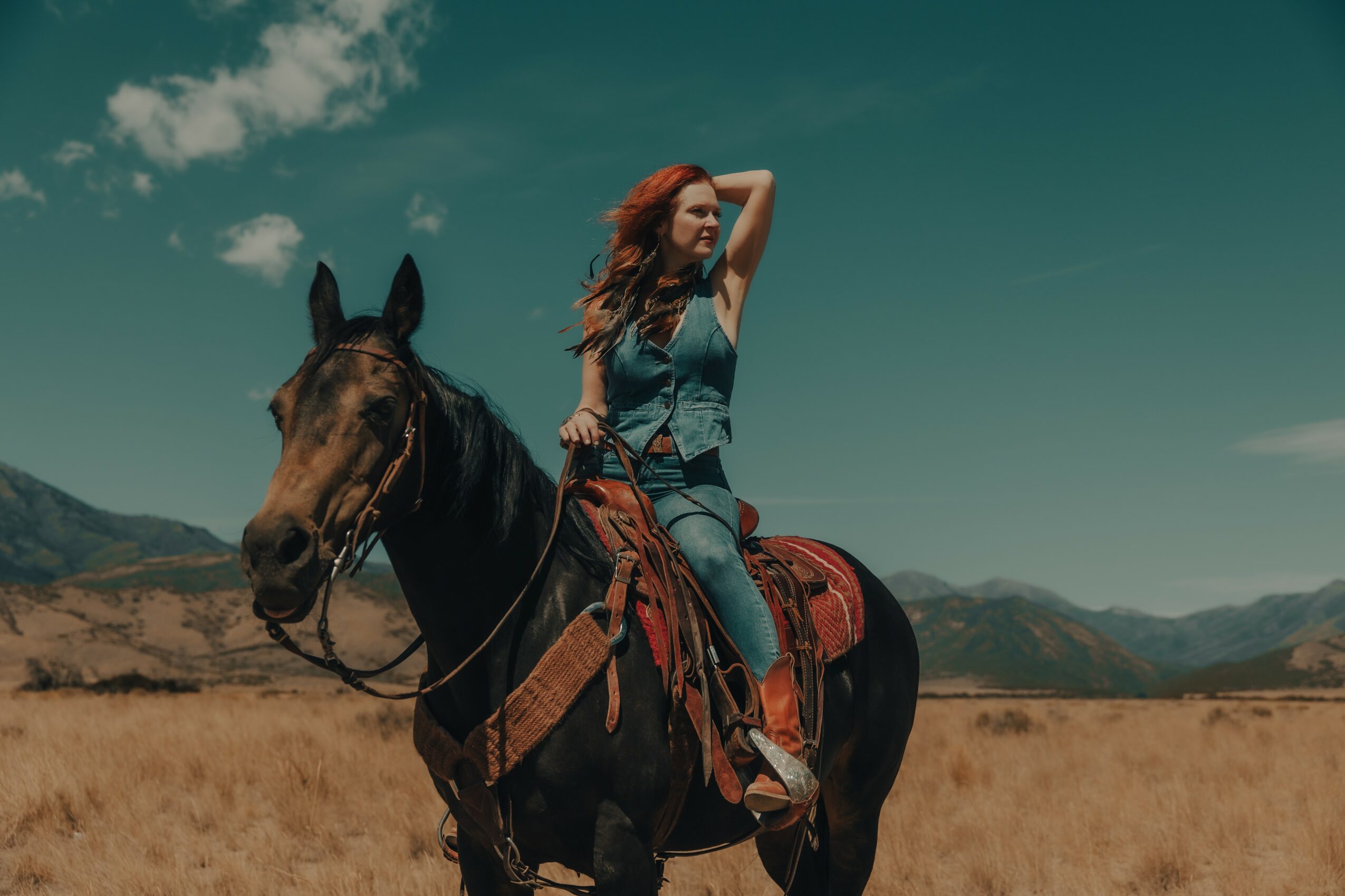 Woman with long hair riding a horse in a vast open field with mountains in the background under a partly cloudy sky.
