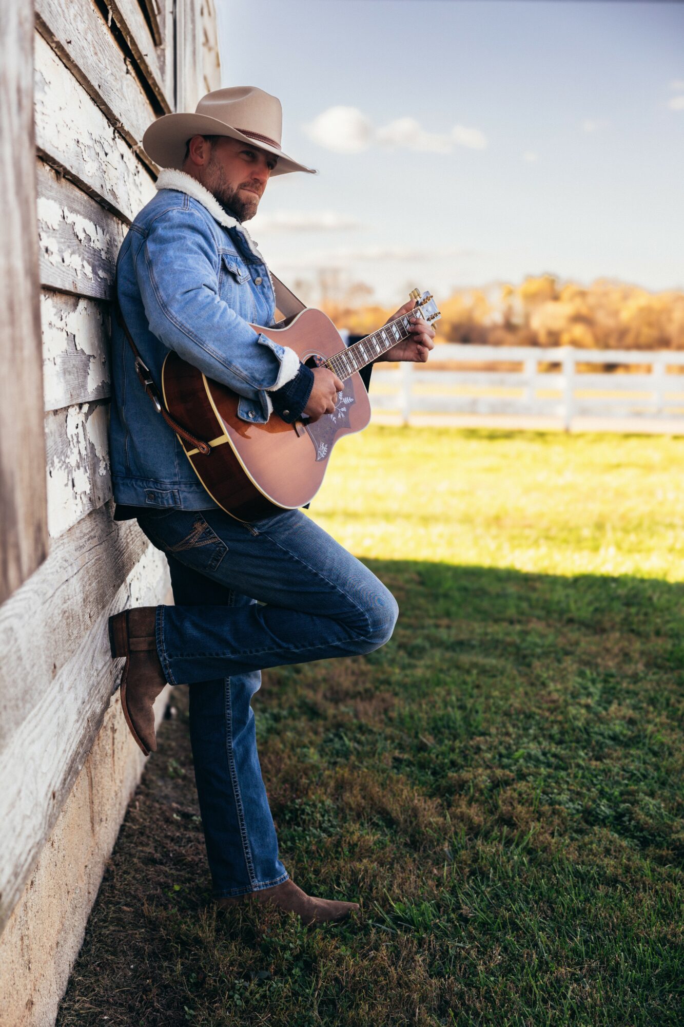 Man wearing a hat and denim jacket playing guitar outdoors, leaning against a wooden wall, with a field and trees in background.