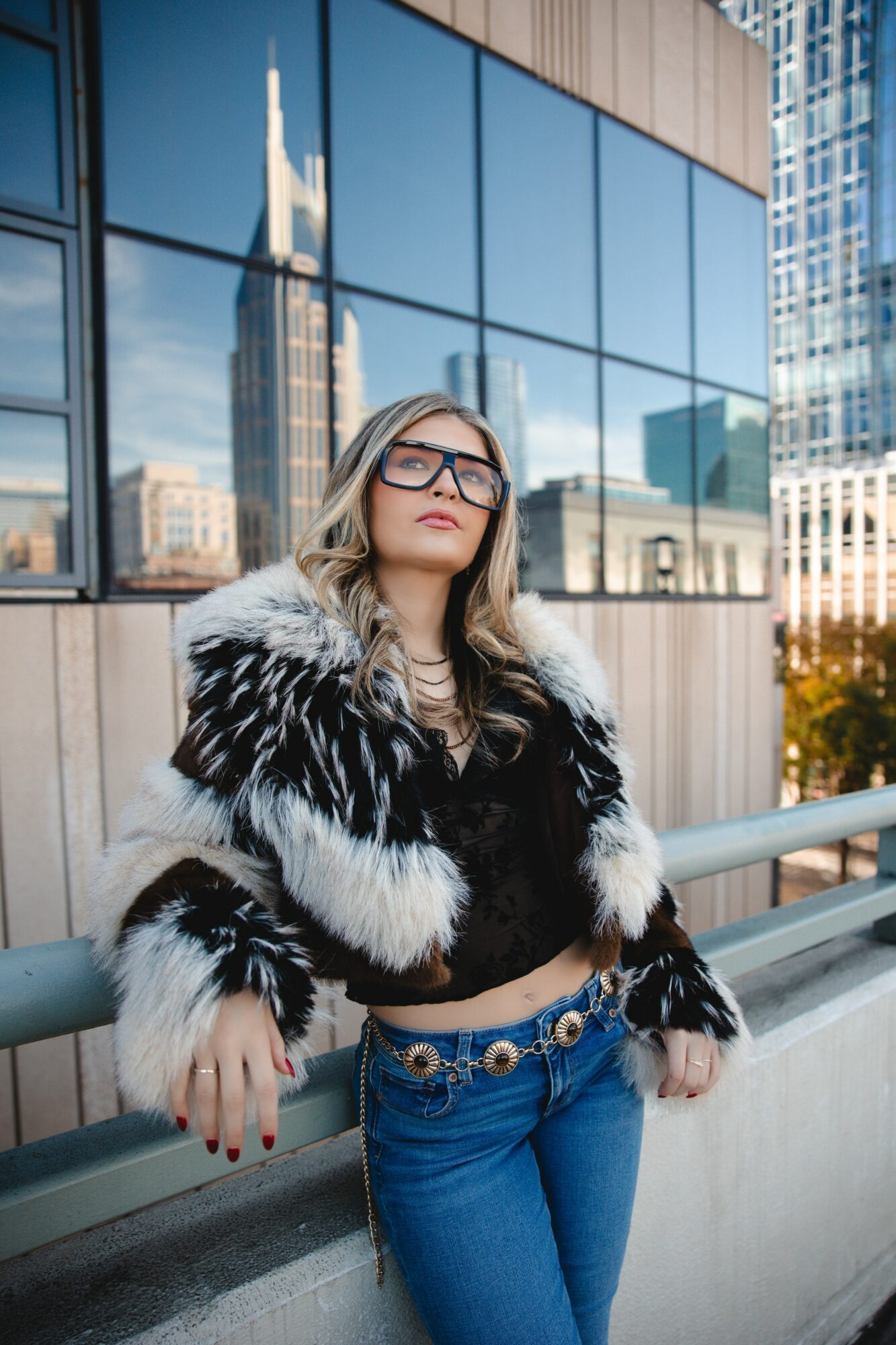 Woman with glasses and long hair in a black and white fur coat, standing outdoors with city buildings reflected in glass windows behind her.
