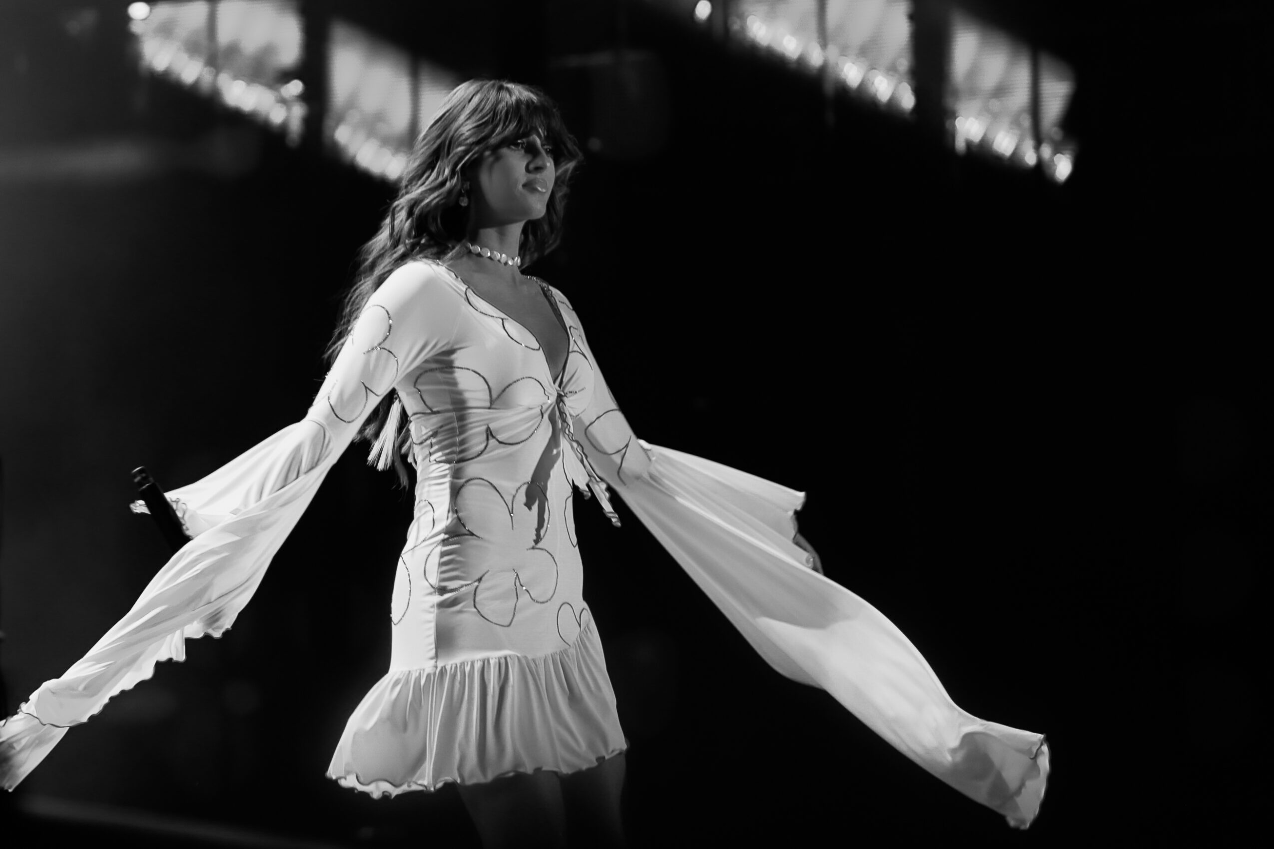 A woman in a dress with long sleeves and ruffled hem stands on stage with arms extended, dark background, black and white.