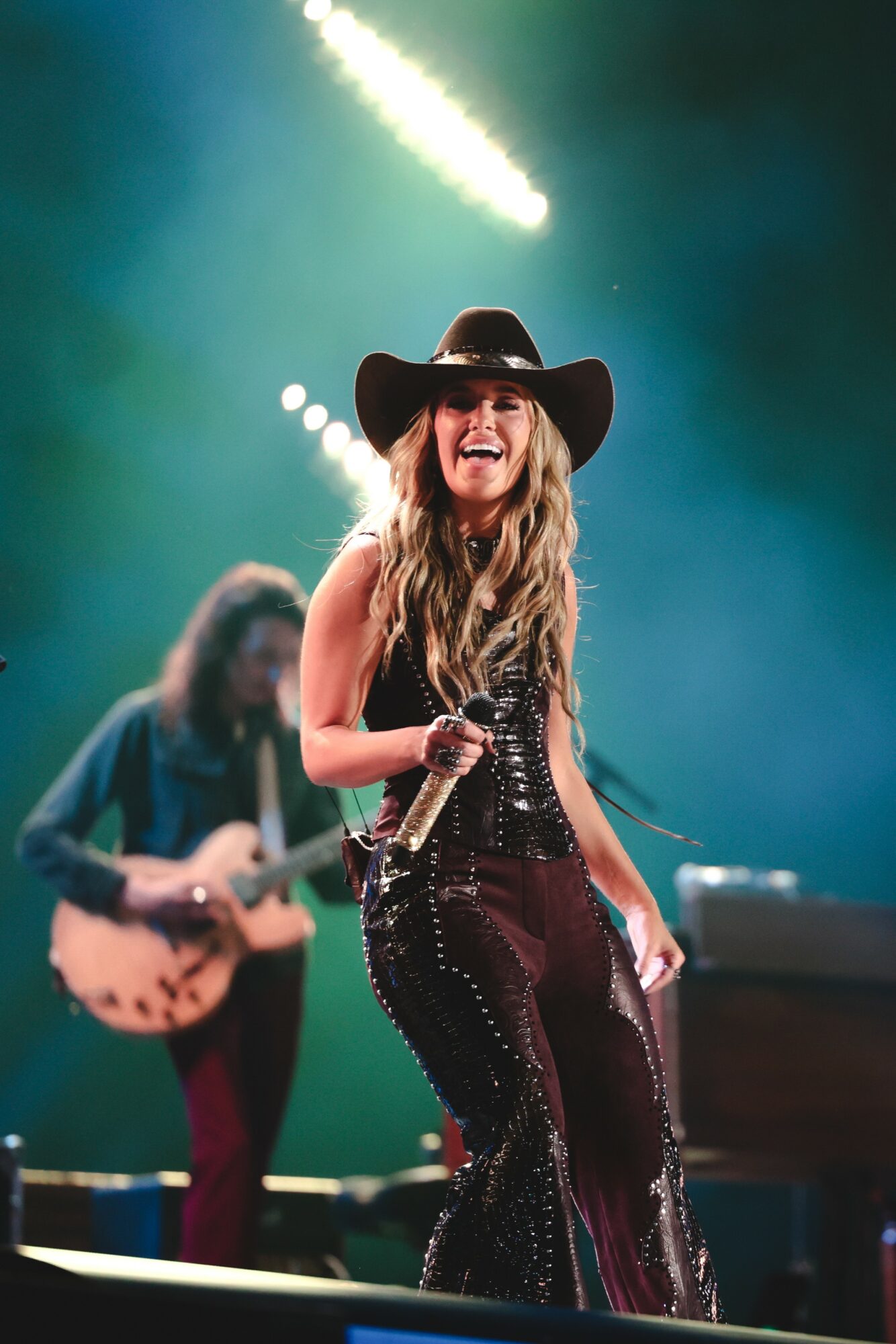 A woman with long hair, wearing a wide-brimmed hat and sequined outfit, singing into a microphone on stage with a guitarist in the background.