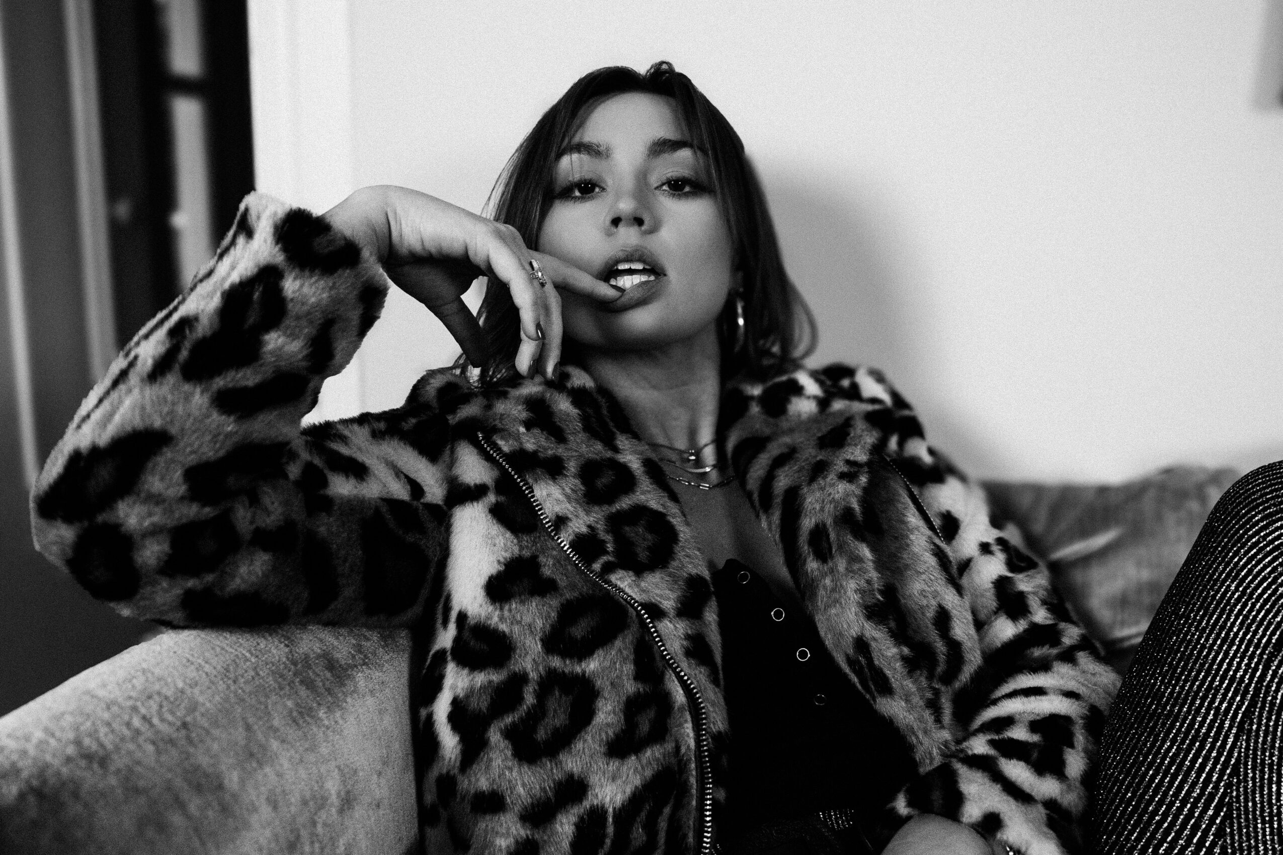 Young woman with long hair wearing a leopard print coat, sitting on a sofa, looking at the camera, black and white photo.