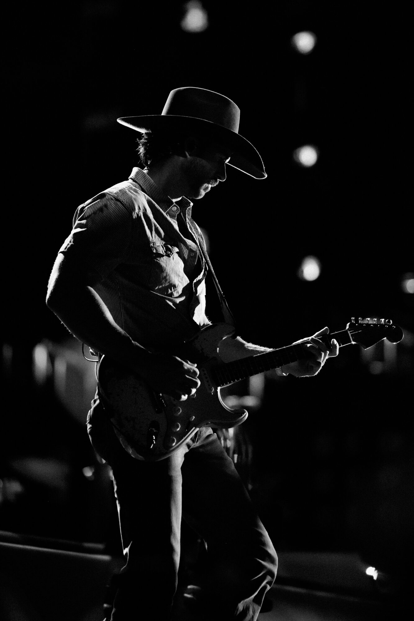 Silhouette of a person playing an electric guitar on stage, wearing a wide-brimmed hat, with stage lights in the background.