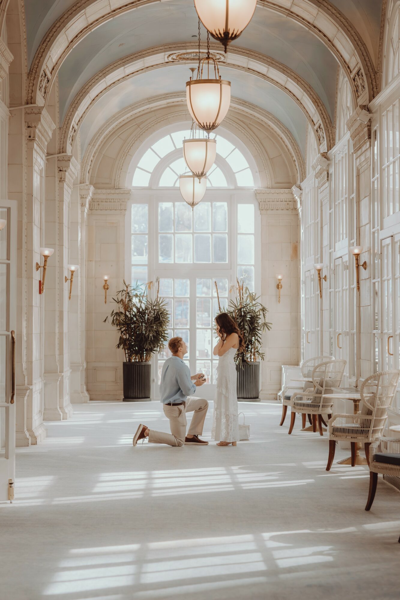 Kneeling person holding hands with standing person in a bright, elegant hallway with large window and chandeliers.