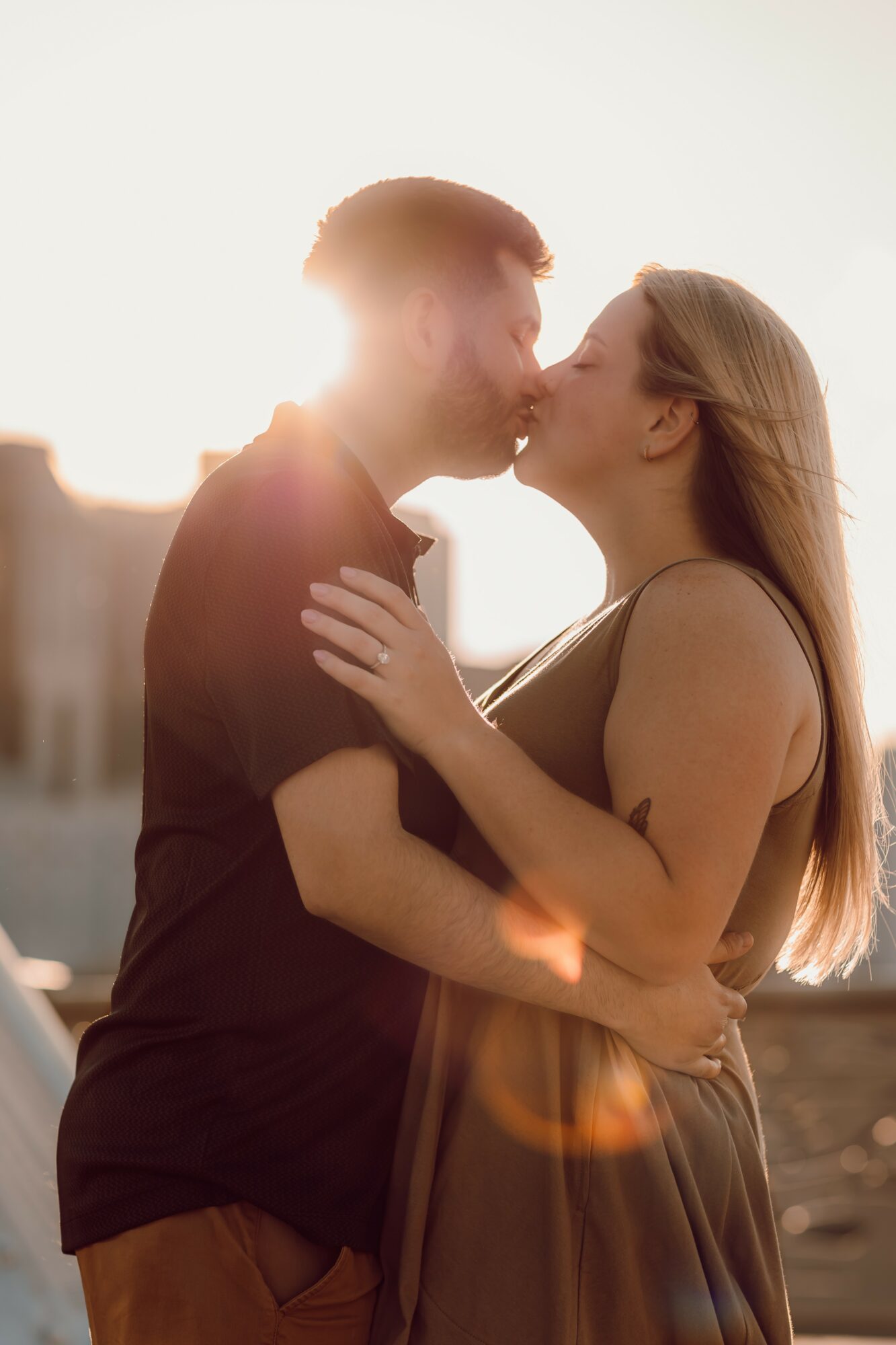 A couple kissing outdoors with sunlight behind them, embracing each other, woman with long hair, man with short hair, warm lighting.