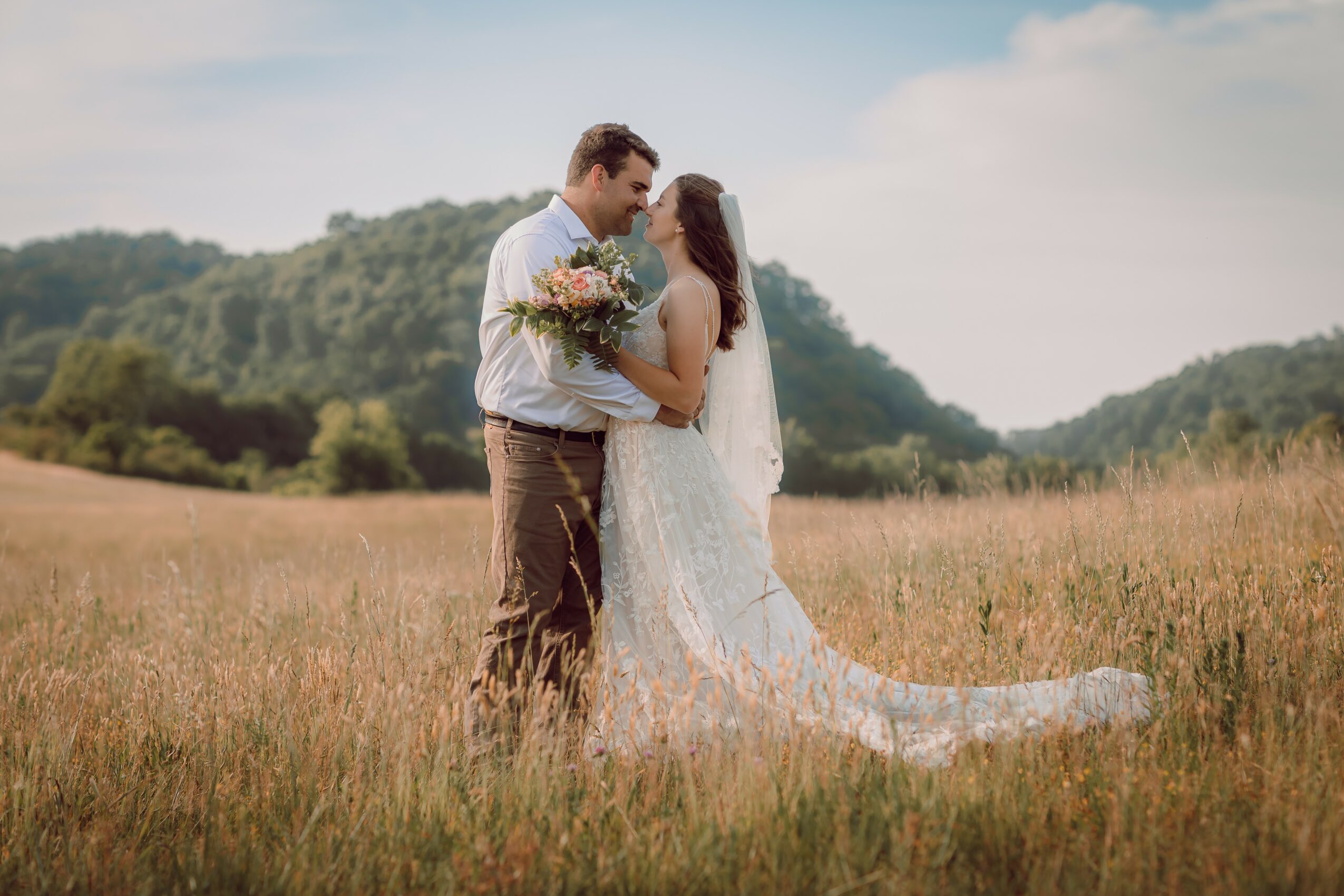 A bride and groom stand close together in a field, holding a bouquet, with hills and sky in the background.