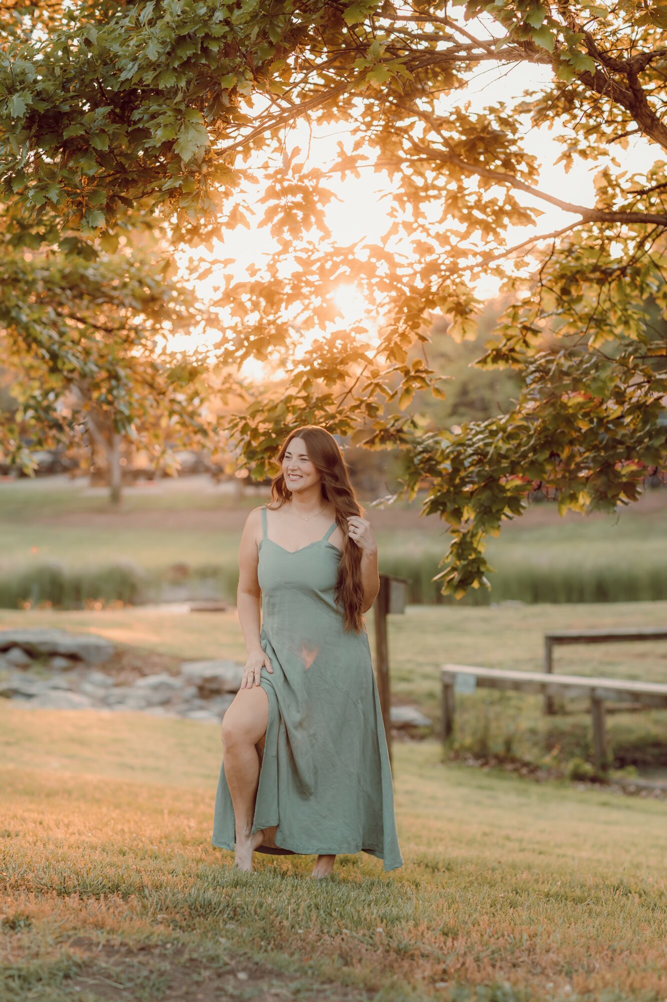 Woman in a long dress standing outdoors near a pond with trees and sunlight in the background.