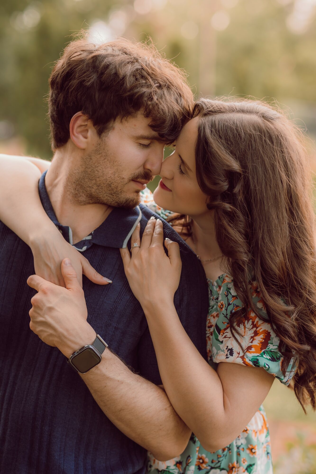 A man and woman face each other closely outdoors, with the woman touching the man's chest and both smiling gently.