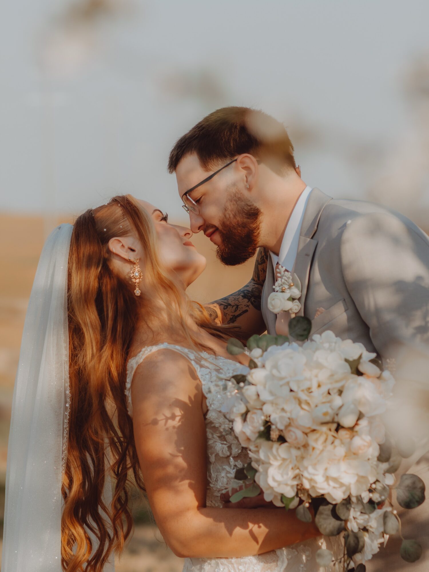 A bride and groom face each other outdoors, smiling, with the bride holding a large bouquet of white flowers.