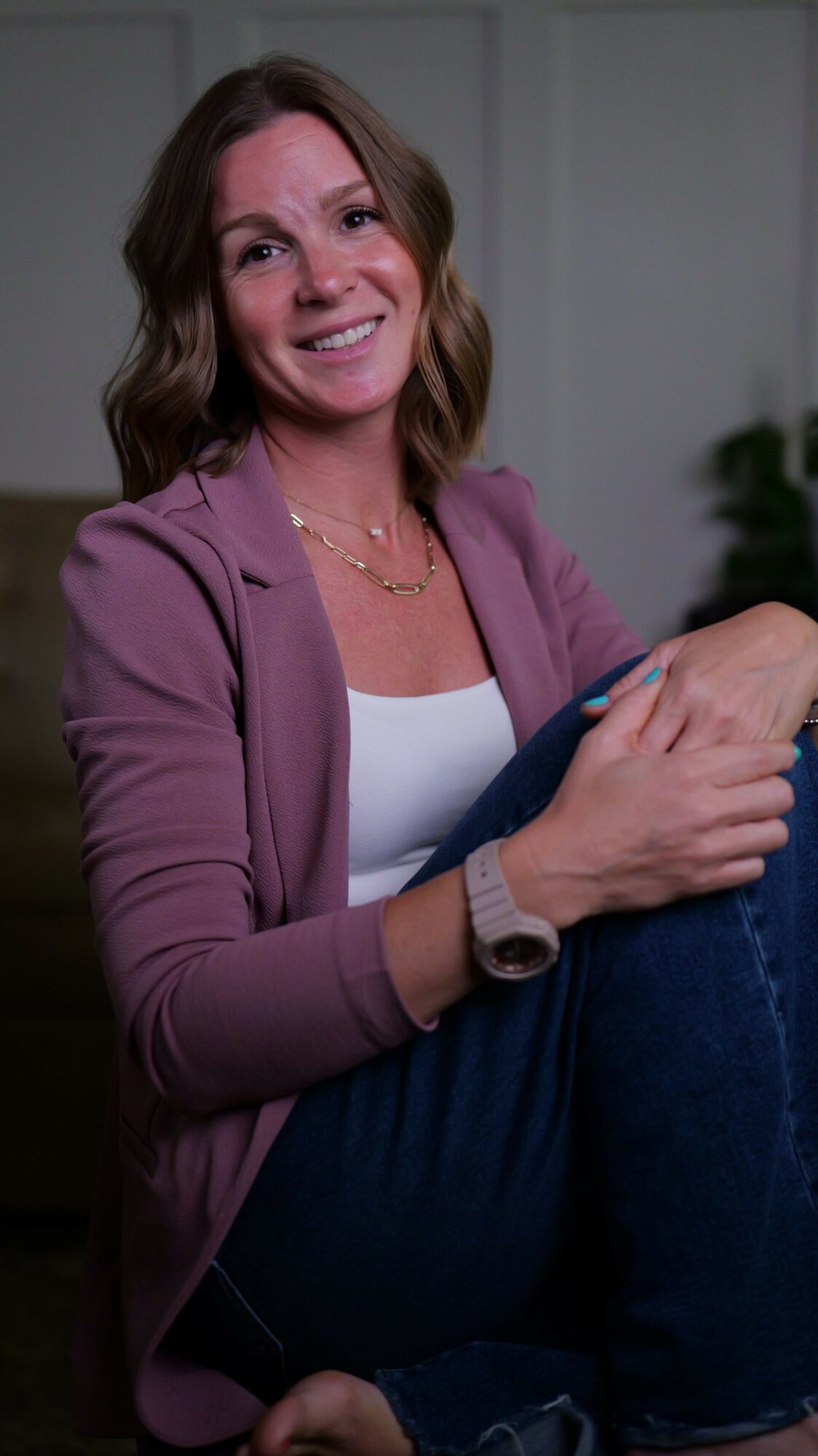 Woman smiling, wearing a pink blazer and white top, sitting with her hands clasped, in a room with neutral background.