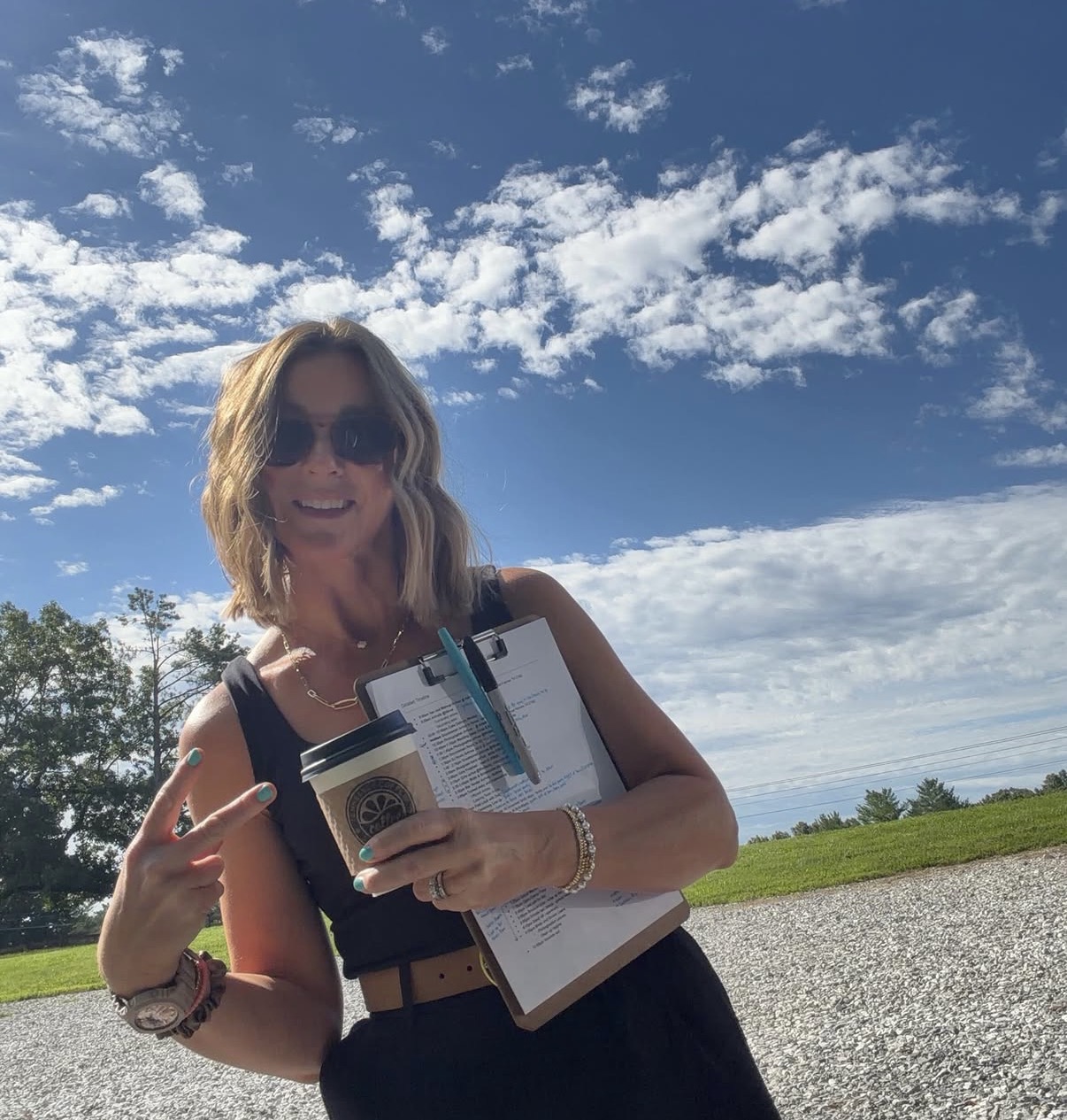 Woman with sunglasses smiling outdoors holding a clipboard and pointing at it, with a blue sky and clouds behind her.