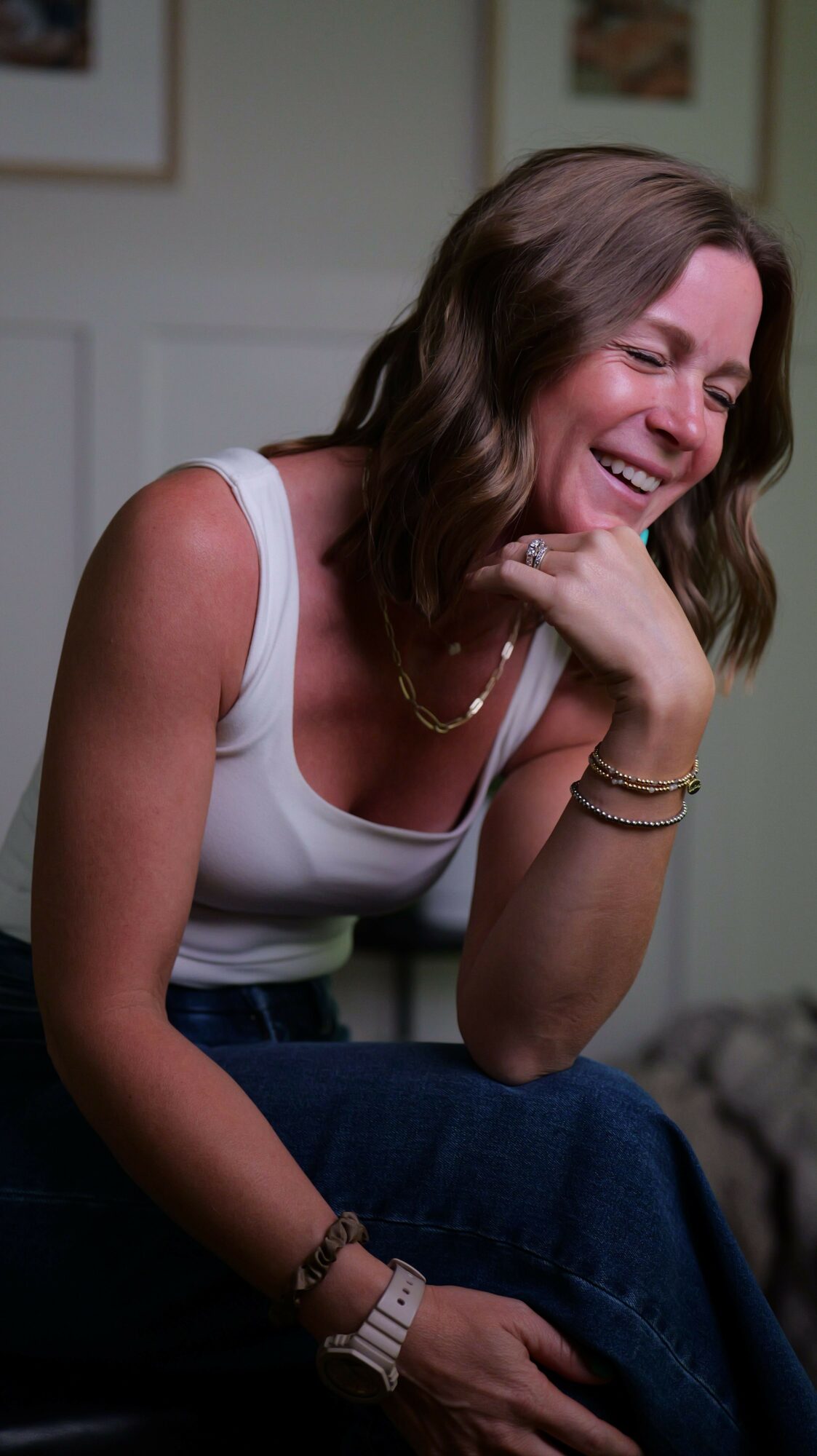 Woman laughing, resting her head on her hand, wearing a white top and jewelry, in a room with framed pictures.