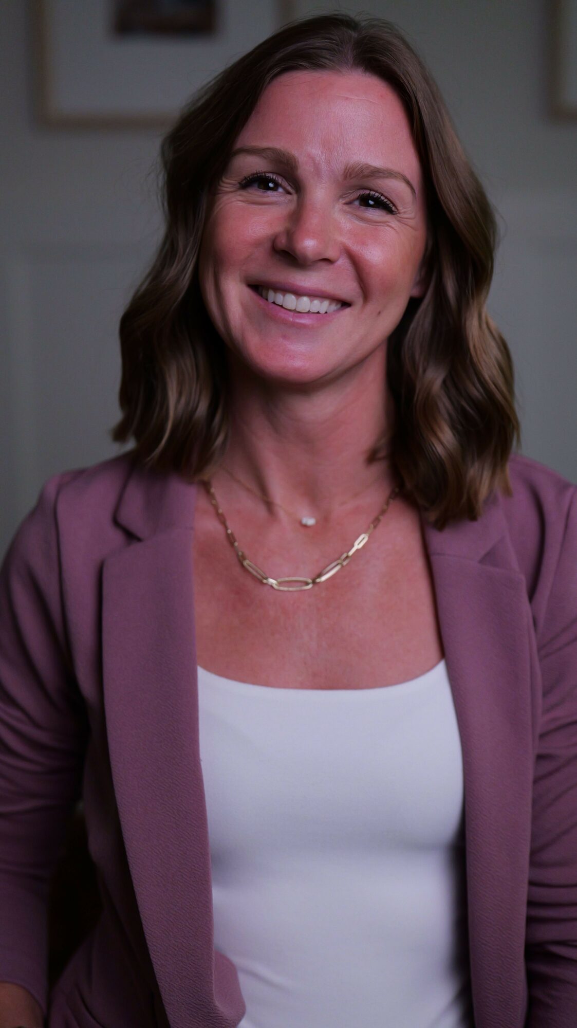 Woman with shoulder-length hair smiling, wearing a white top and purple jacket, in an indoor setting.