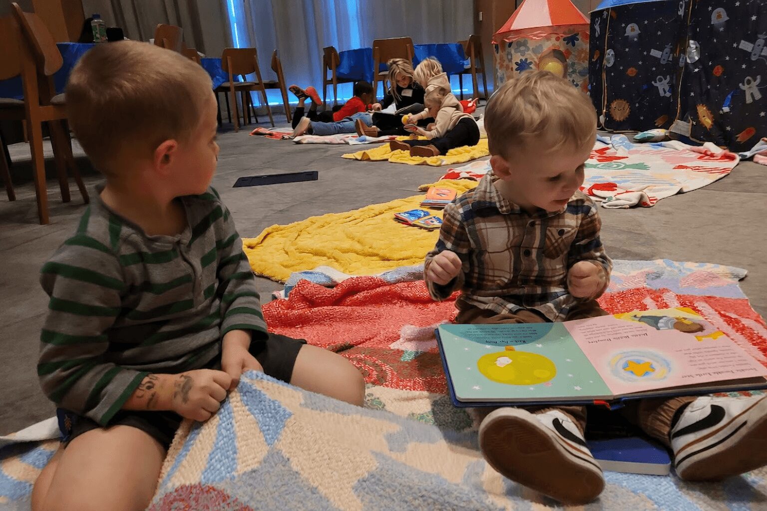 Two young children sitting on a colorful blanket, looking at a picture book, with other children and blankets in the background.