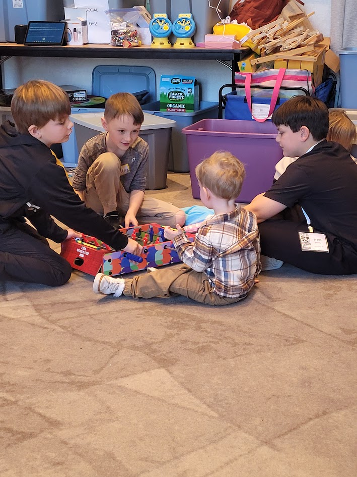 Four children and two adults sit on the floor playing with a toy train, in a room with shelves and storage bins.