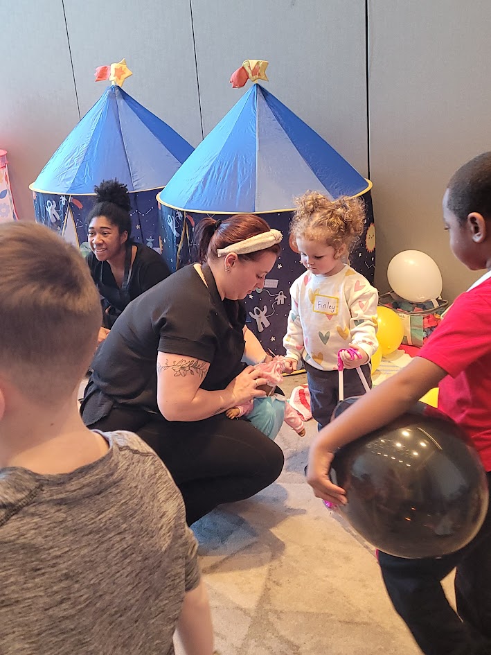 Adult woman interacting with children in front of blue tents, one child holding a black balloon, indoor setting.