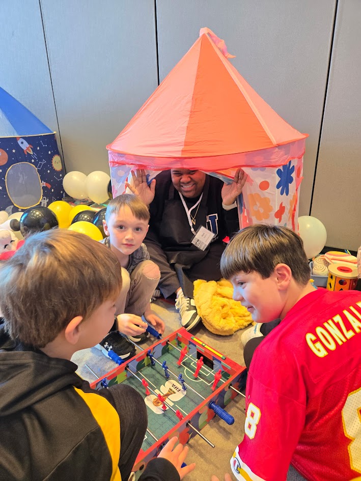 Children and an adult playing foosball under a pink and orange tent at a party or event.
