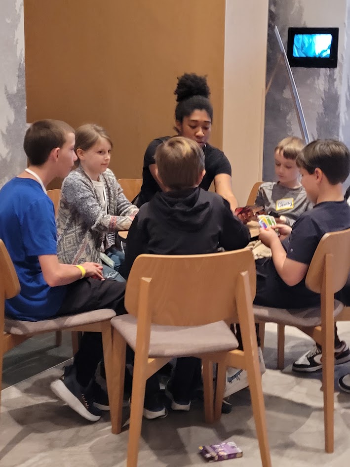 Group of six children and one adult sitting around a table, engaging in a game or activity, in a room with beige and gray walls.