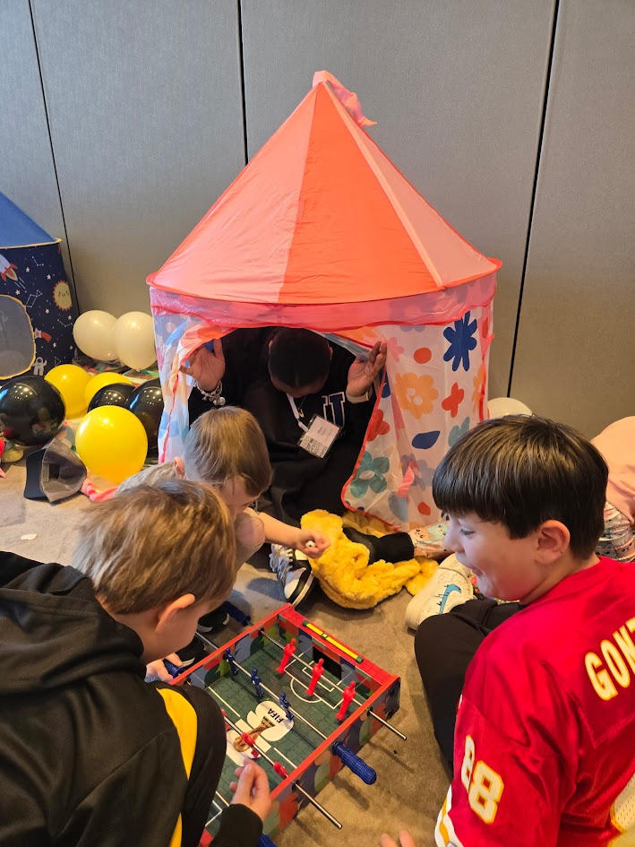 Children gather around a small pink and orange play tent with colorful flower patterns, engaging with toys inside.
