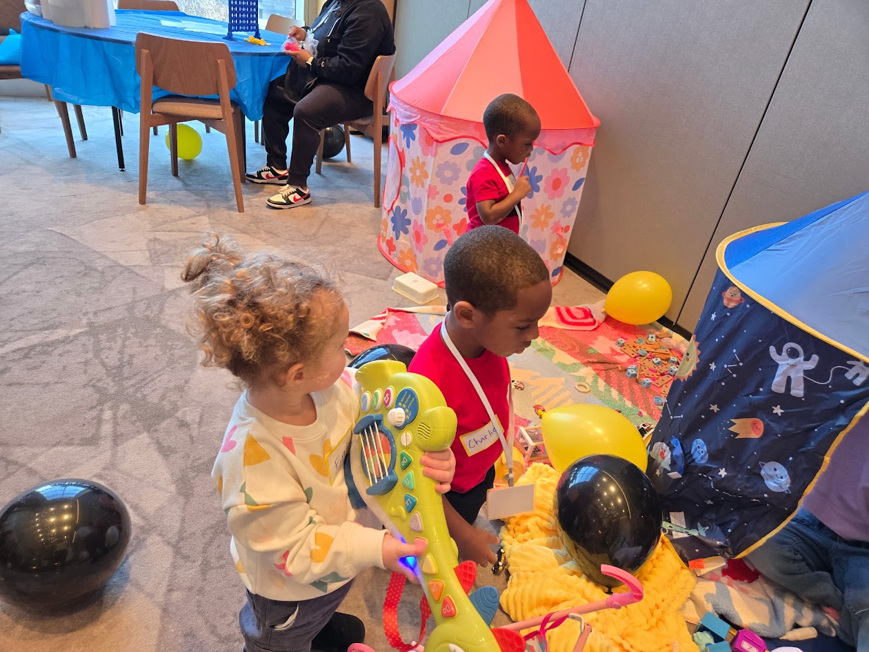 Children playing with balloons and toys indoors, with a pink play tent and a blue play area in the background.