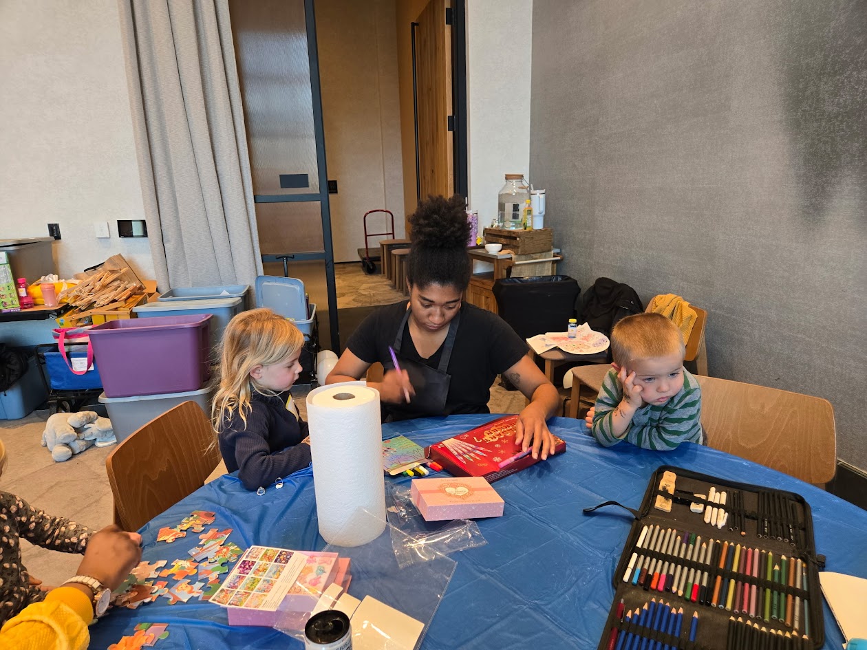 Woman with two children sitting at a table with art supplies and paper towels, in a room with a gray wall.