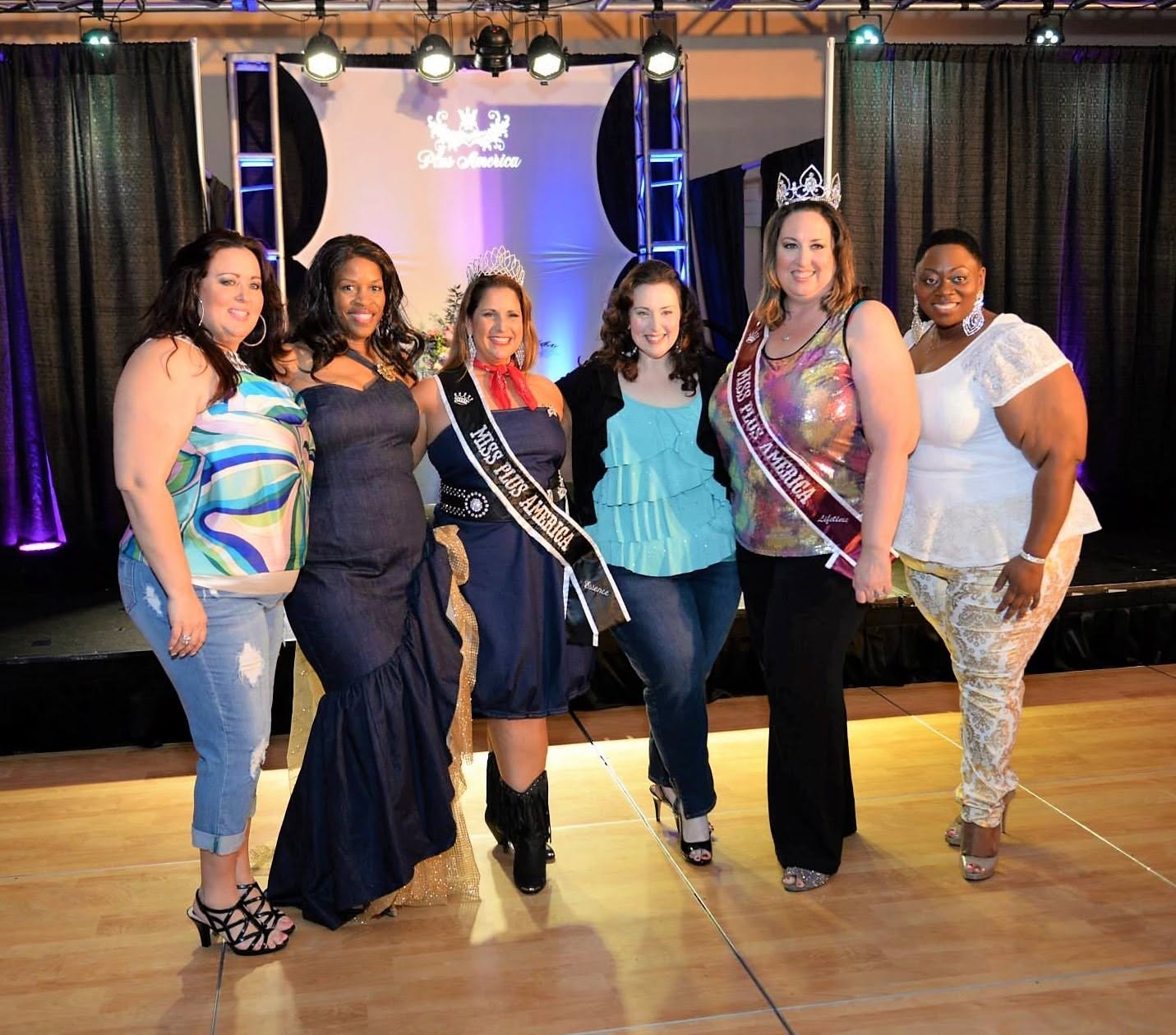 Six women standing on a stage with a black curtain and purple lighting, smiling for a group photo.