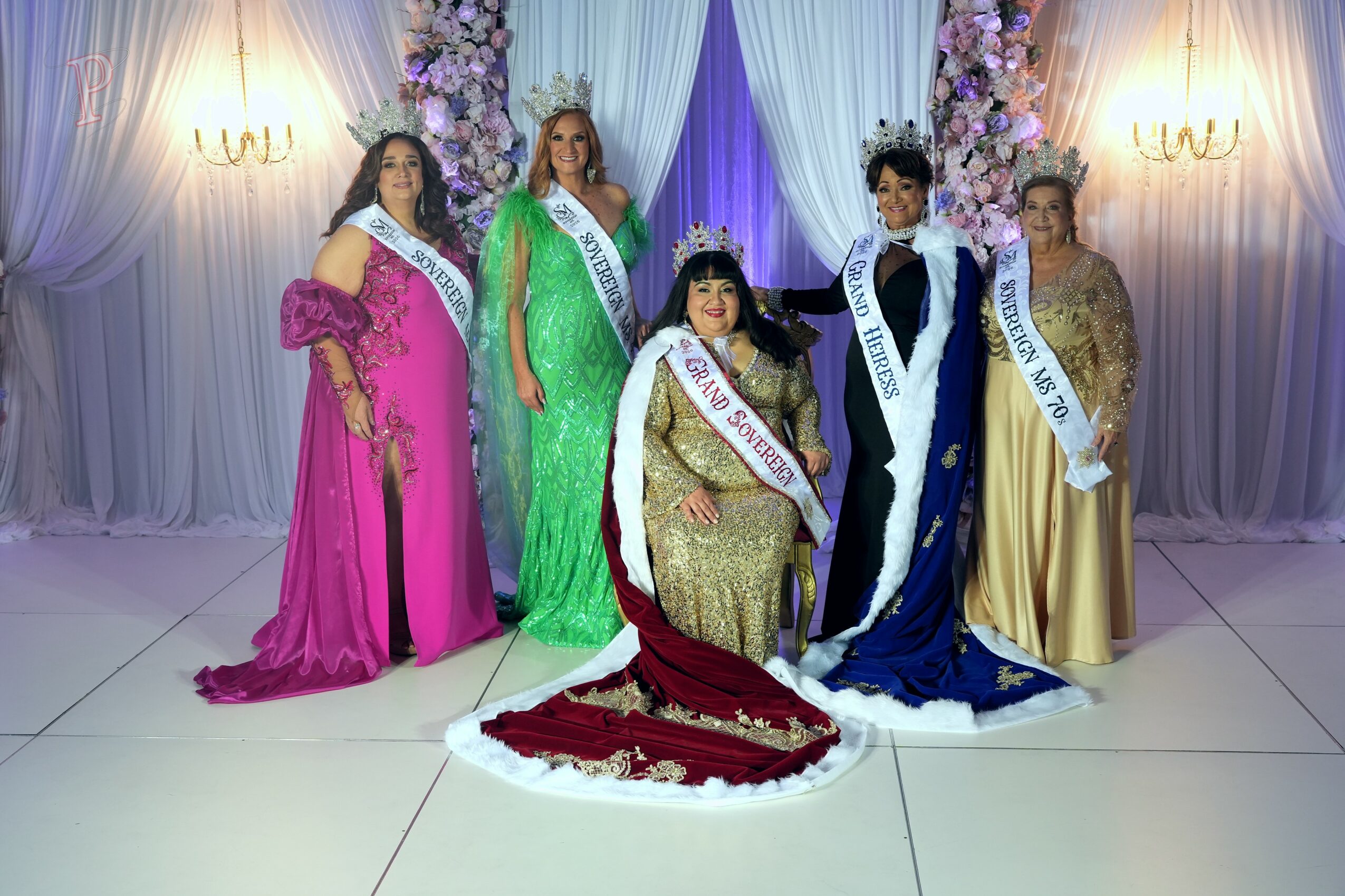 Six women in colorful gowns and sashes pose in front of floral decorations and draped curtains, with two wall sconces on each side.