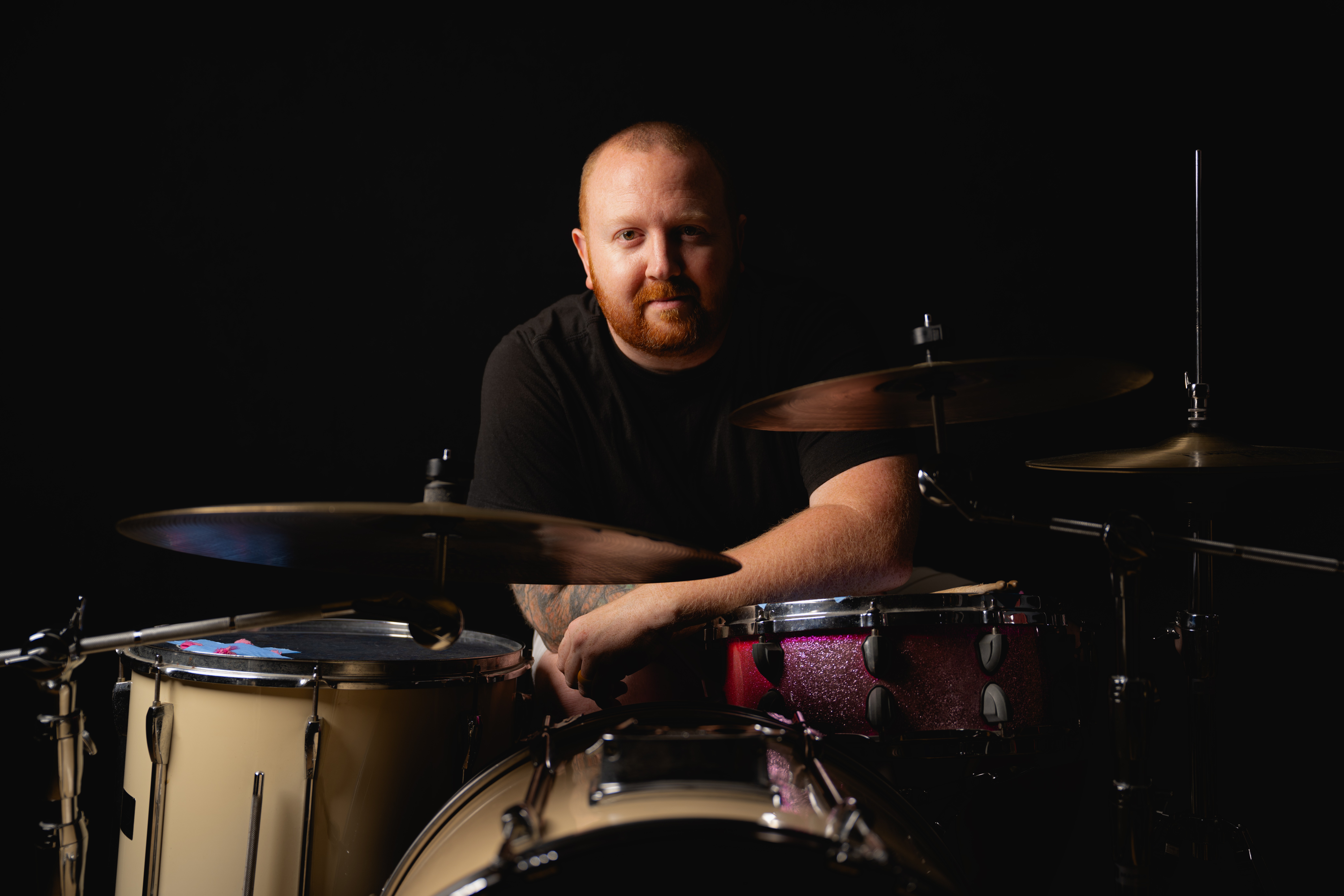 Man with a beard sitting behind a drum set in a dark background.