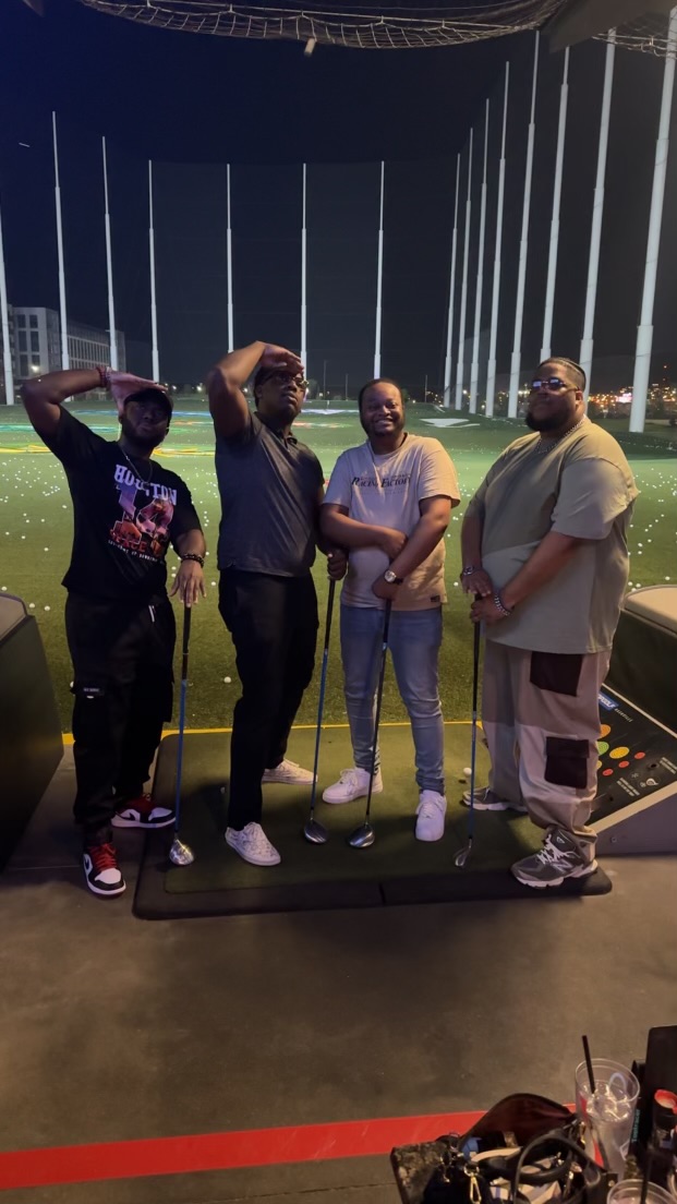 Four people standing together outdoors at night, with a tall fence and lights in the background.