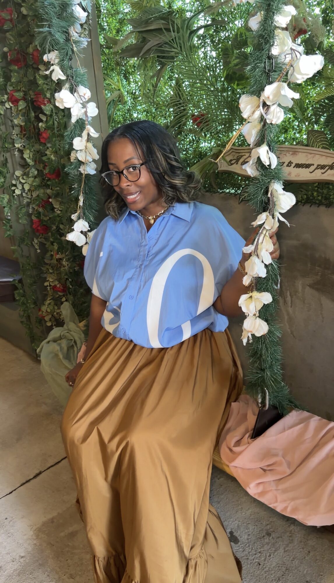 Woman sitting in front of floral backdrop, wearing glasses, blue top, and brown skirt, smiling at camera.