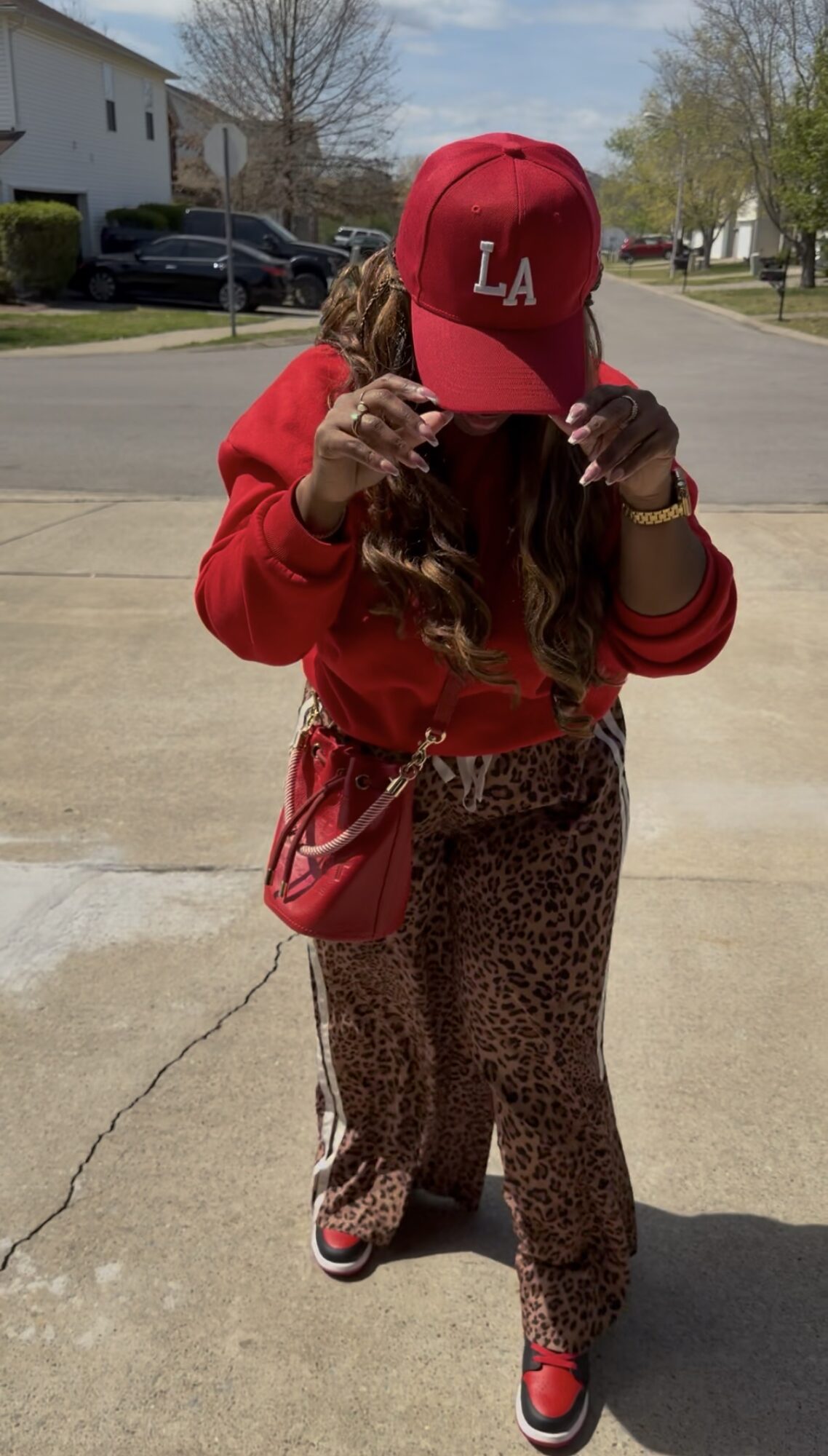 Person wearing a red LA cap, red sweatshirt, leopard print pants, and red shoes, standing outdoors on a sunny day.