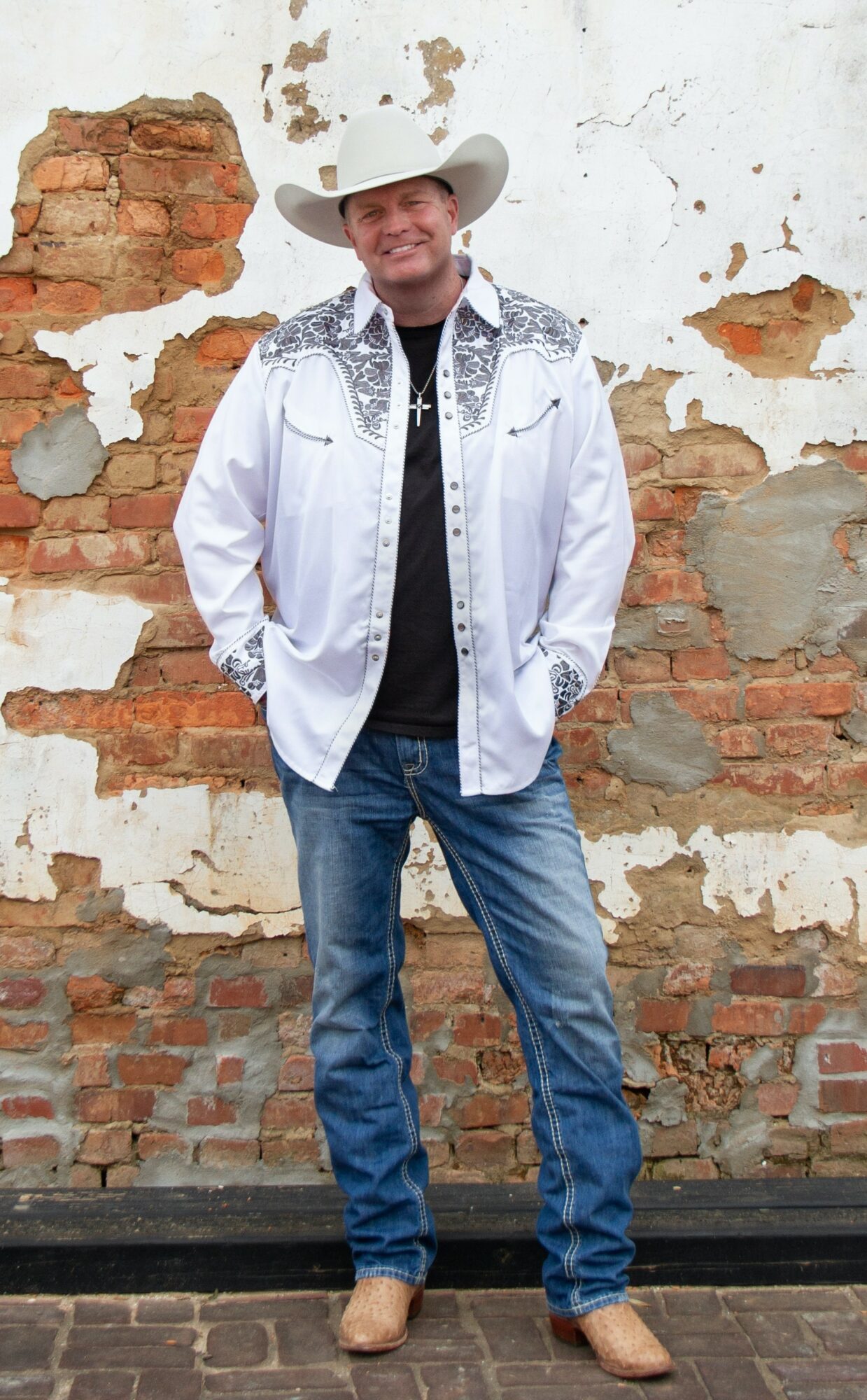 Man in cowboy hat, white jacket, black shirt, jeans, standing outdoors against a weathered brick wall.