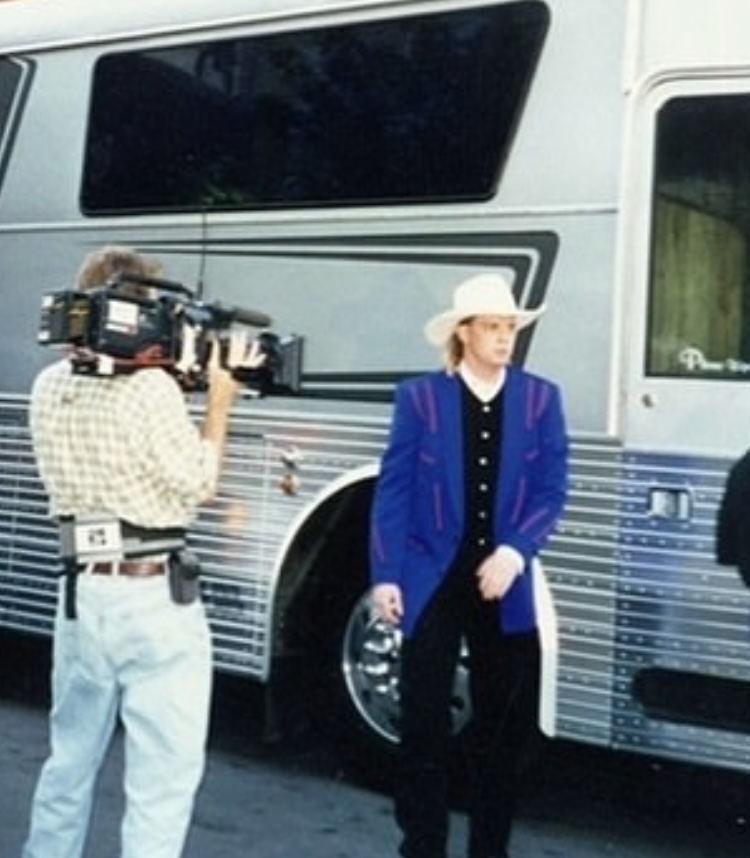 Person with a camera filming a woman in a blue blazer and white hat near a bus.