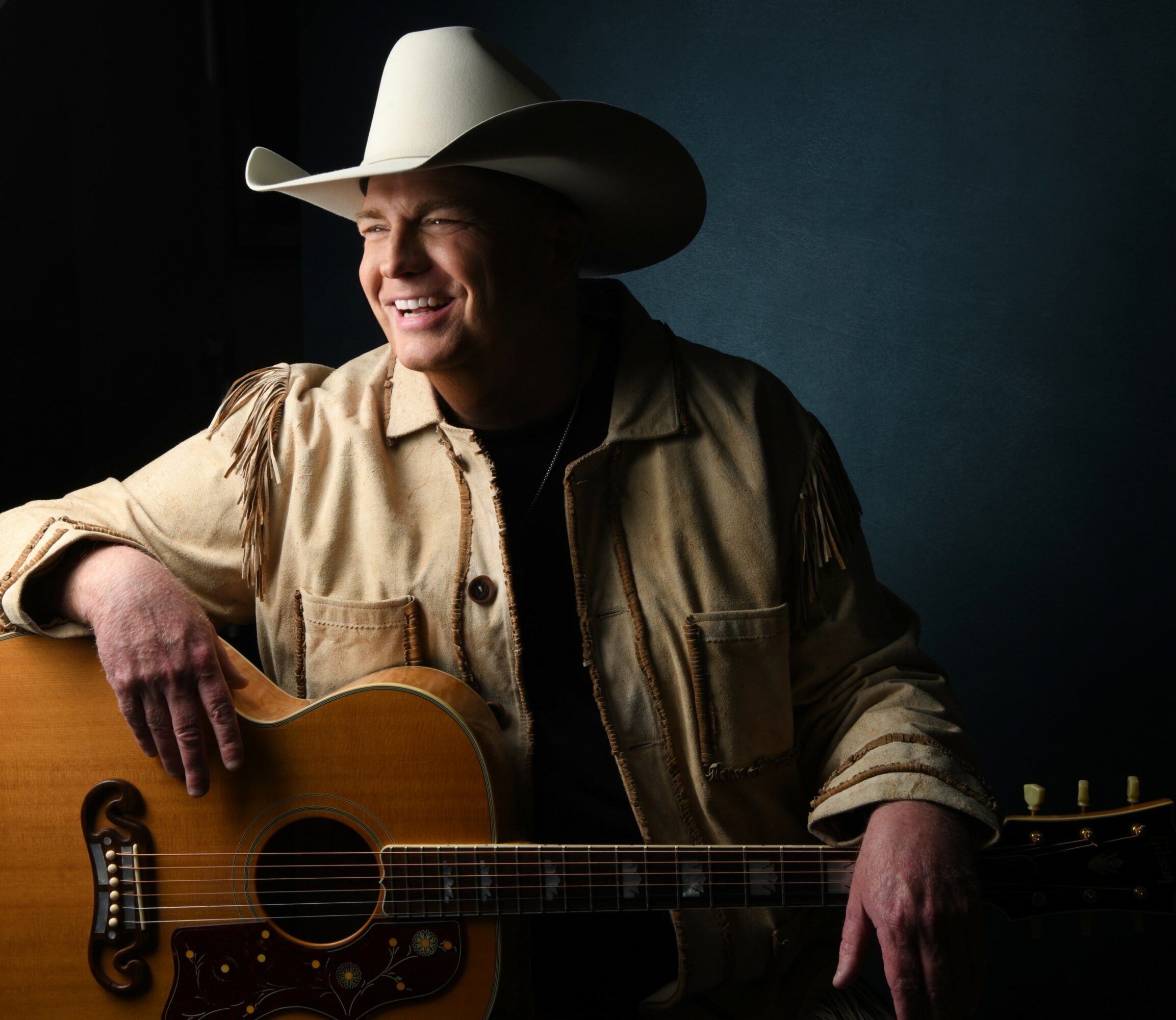 Person wearing a cowboy hat and beige jacket, smiling, holding an acoustic guitar, against a dark background.