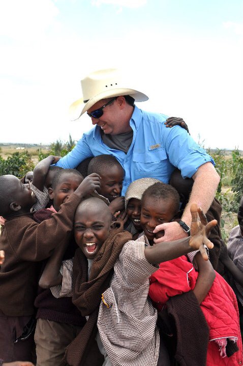 Woman in hat and sunglasses surrounded by children outdoors, smiling and interacting with them.