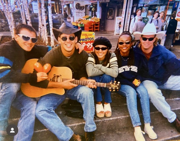Group of five people sitting on a bench outdoors, smiling, with one person holding a guitar, all wearing sunglasses.