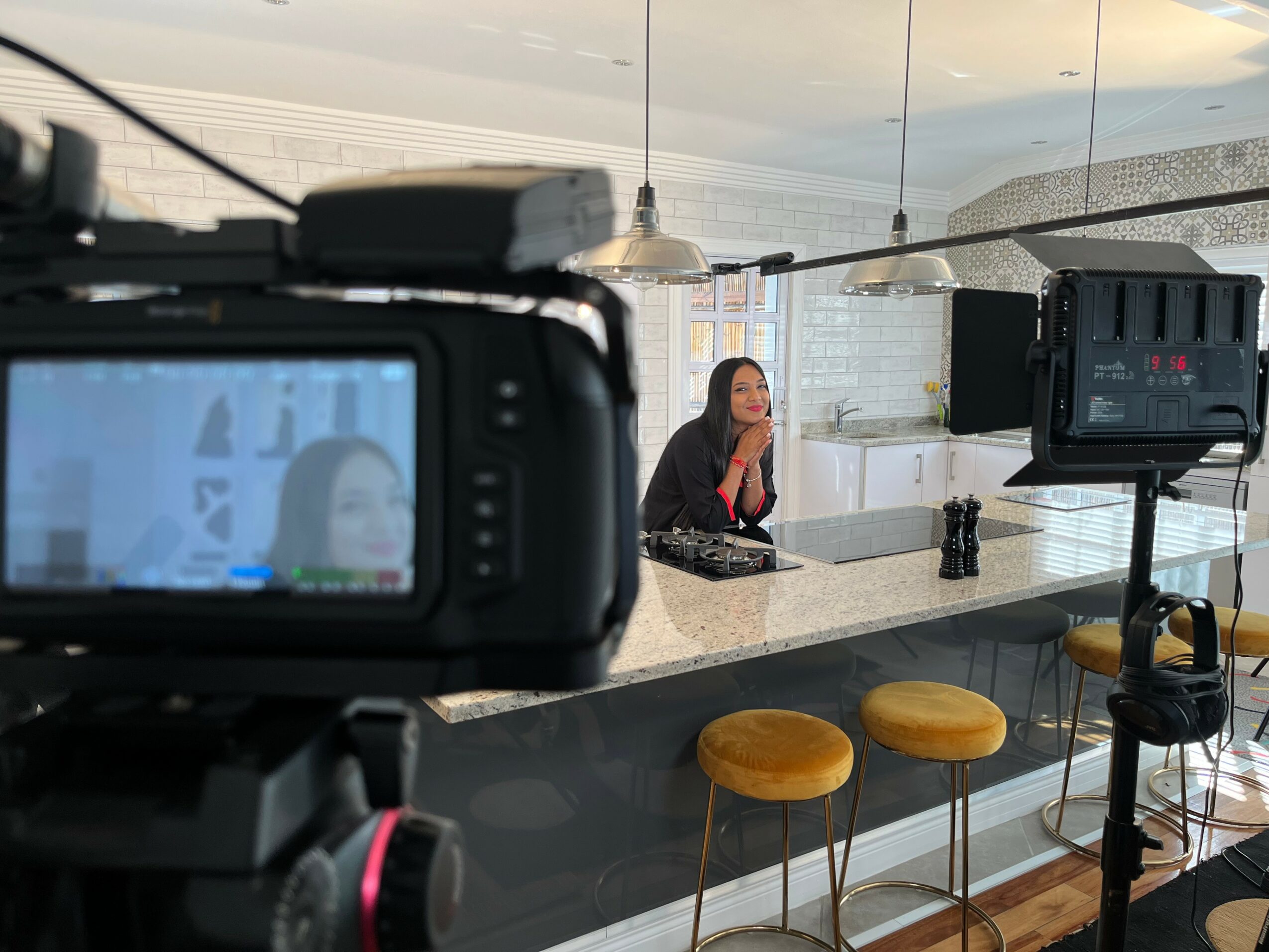 A woman sitting at a counter in a modern kitchen, with a camera recording her, and three yellow stools in front.