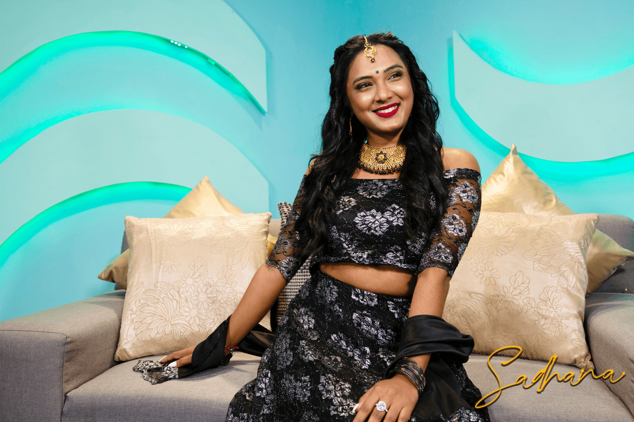 Woman in black floral outfit smiling, sitting on a sofa with beige pillows, colorful background, jewelry, and makeup.