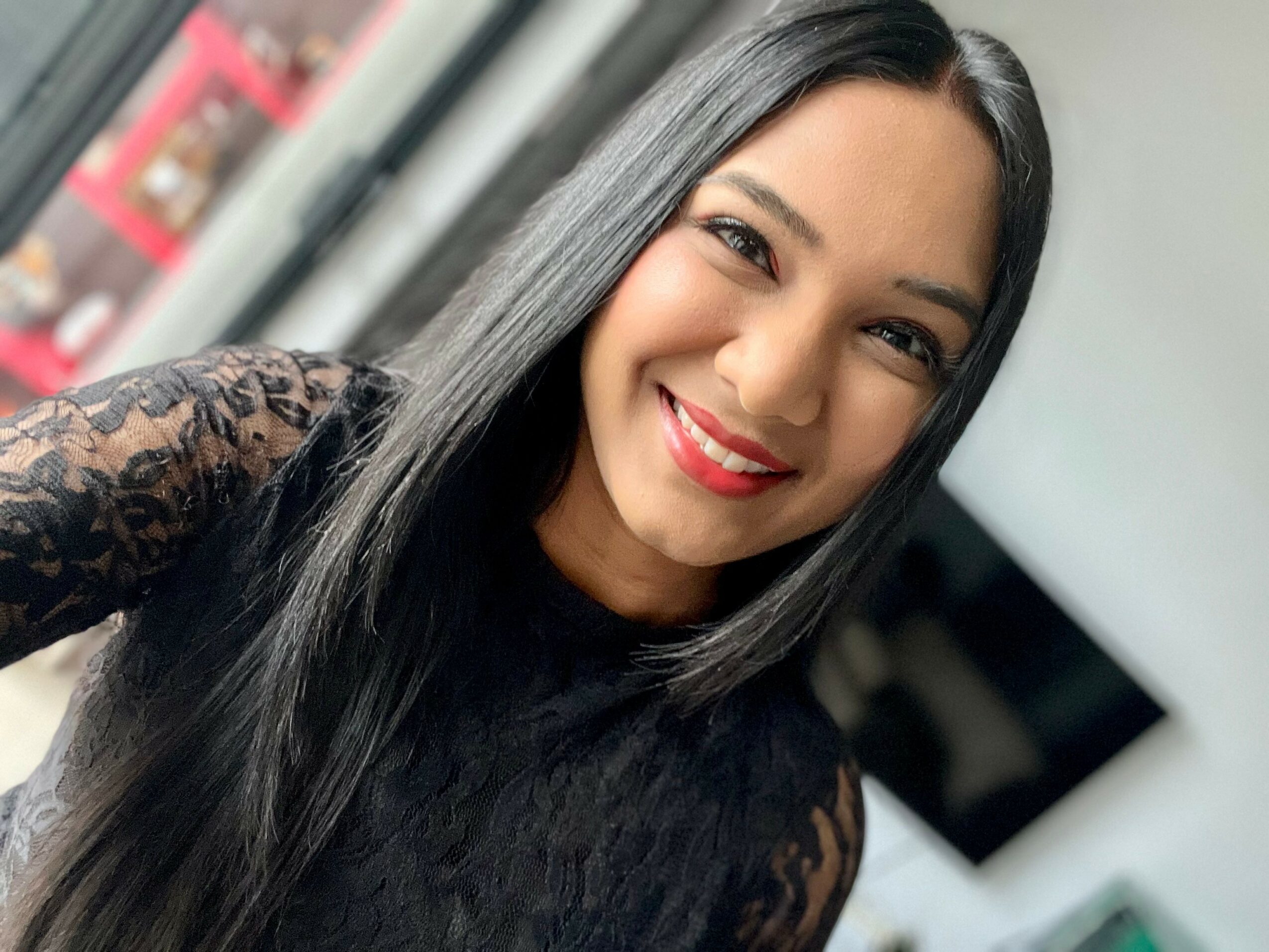Smiling woman with long black hair wearing a black lace top, indoors near a window.