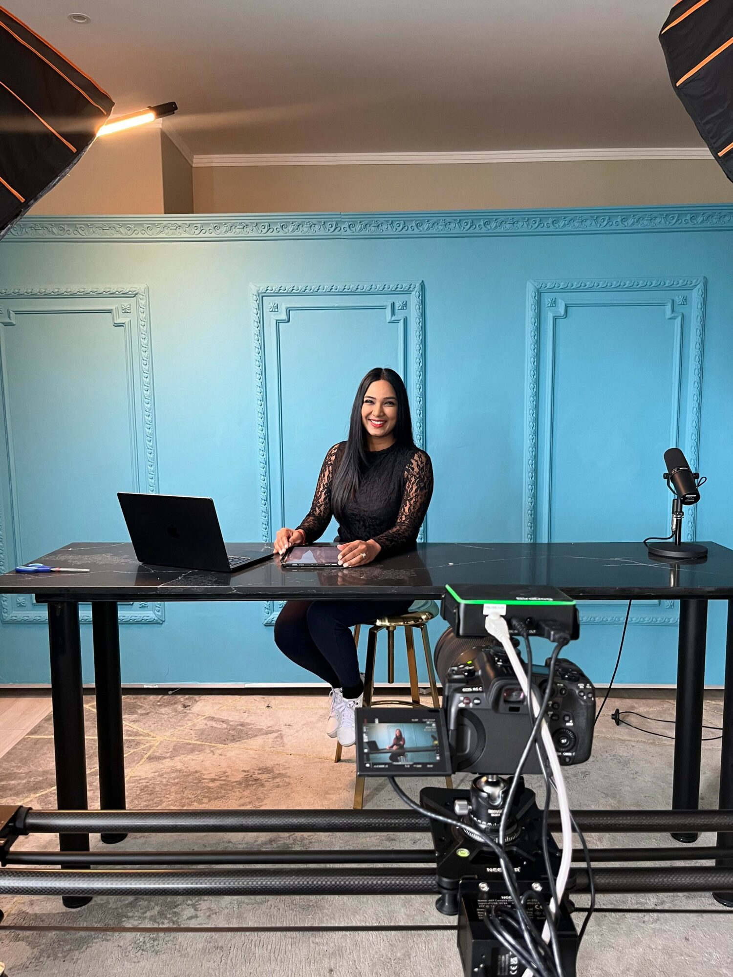 Woman standing behind a desk with a laptop, in a room with blue walls and decorative molding.