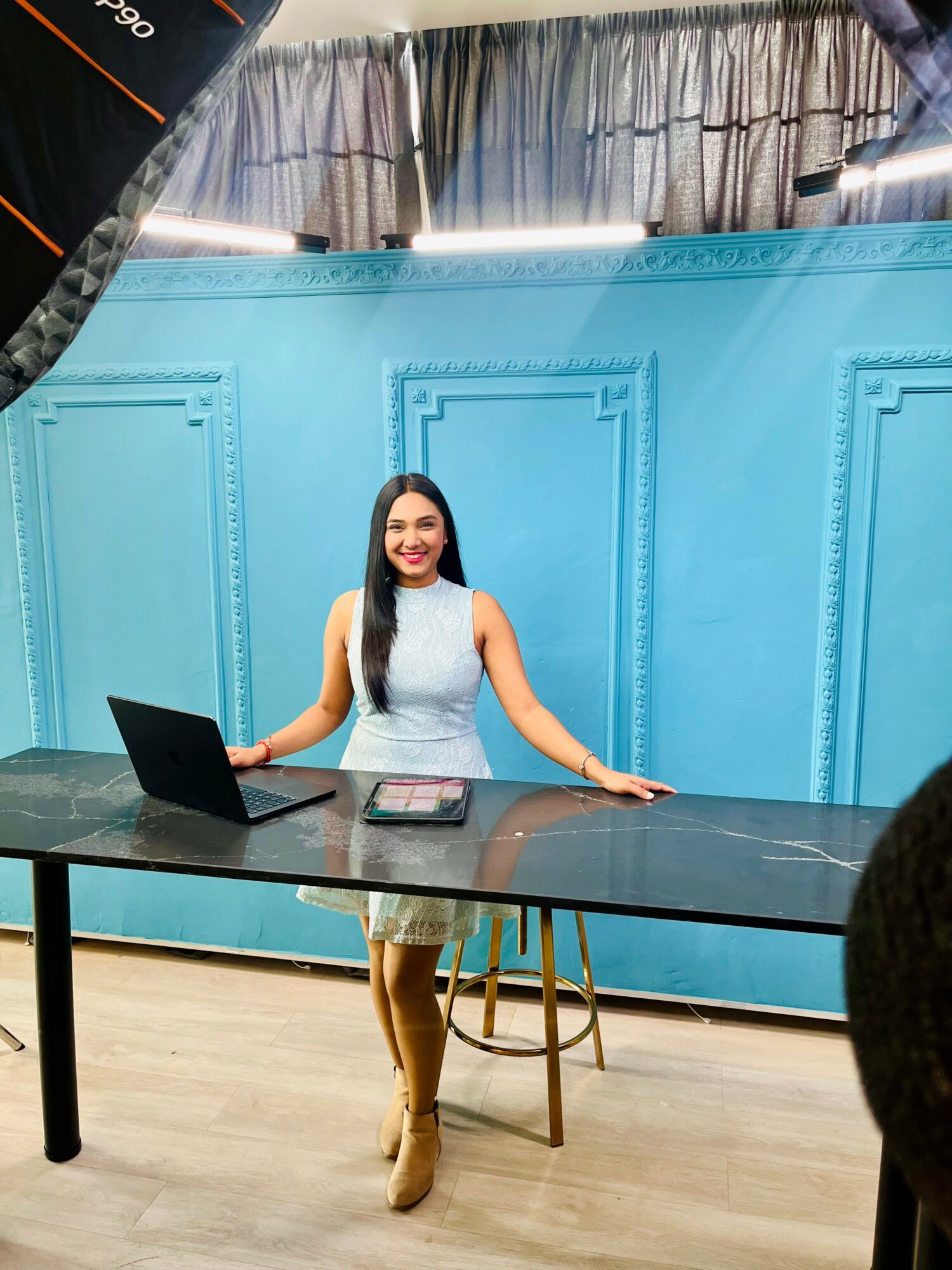 Woman sitting at a desk with a laptop, smiling, in front of a blue backdrop, with a curtain above.